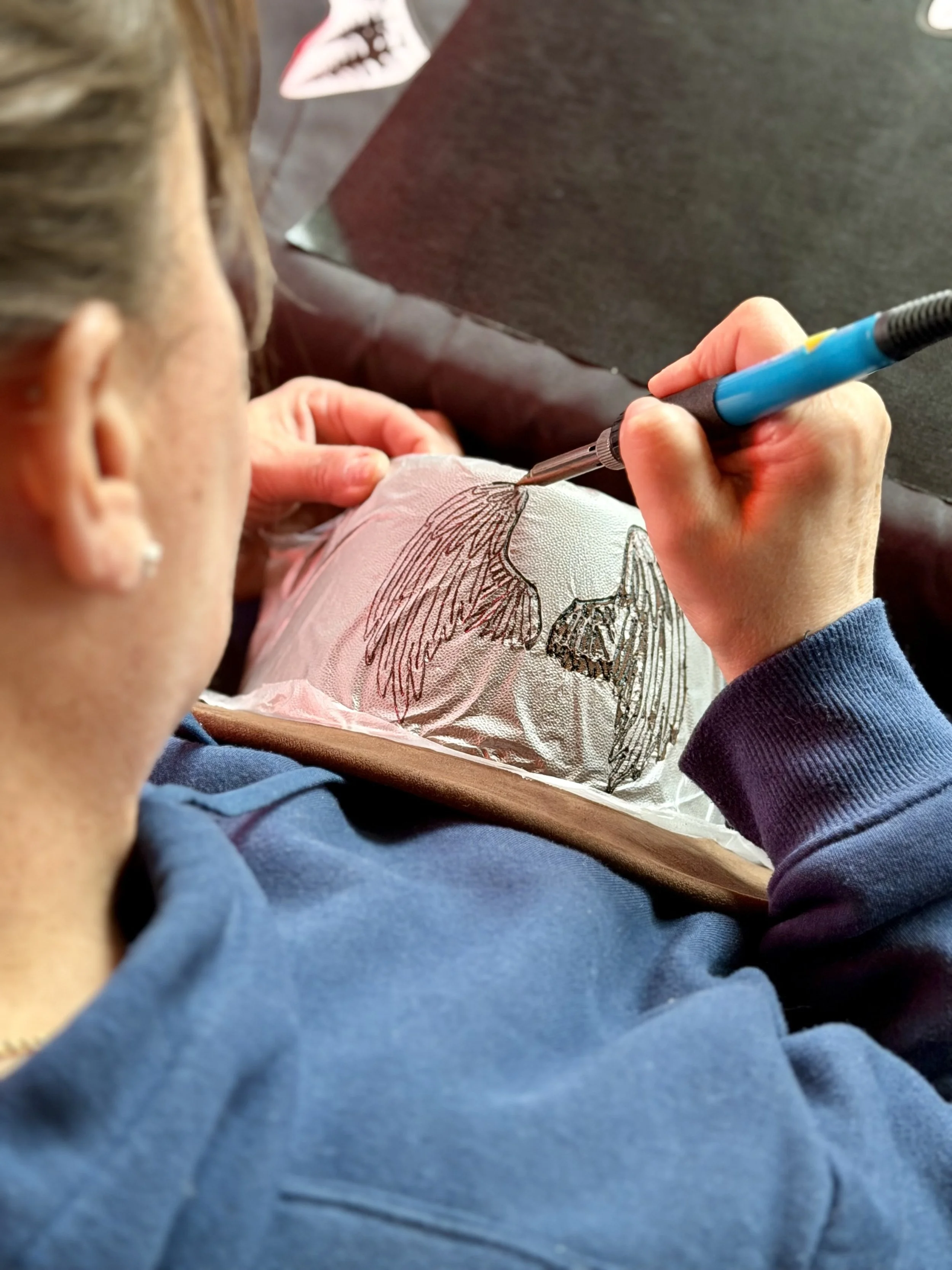Close-up of a person in a blue shirt, holding a pen, drawing a detailed bird illustration on a piece of paper on their lap, with another person wearing a purple shirt near them.