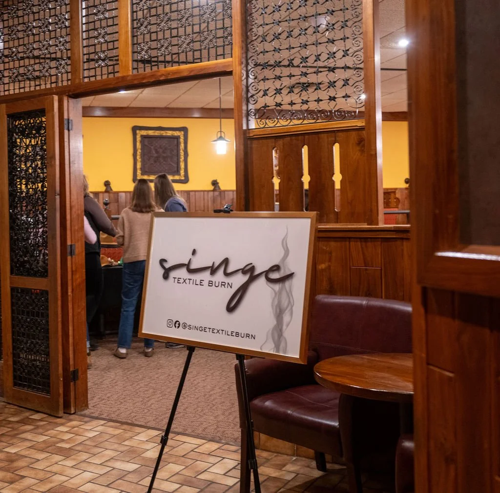 A restaurant interior with wooden decor, a signboard that reads 'Single Textile Burn' with social media handles, and a group of people in the background talking.