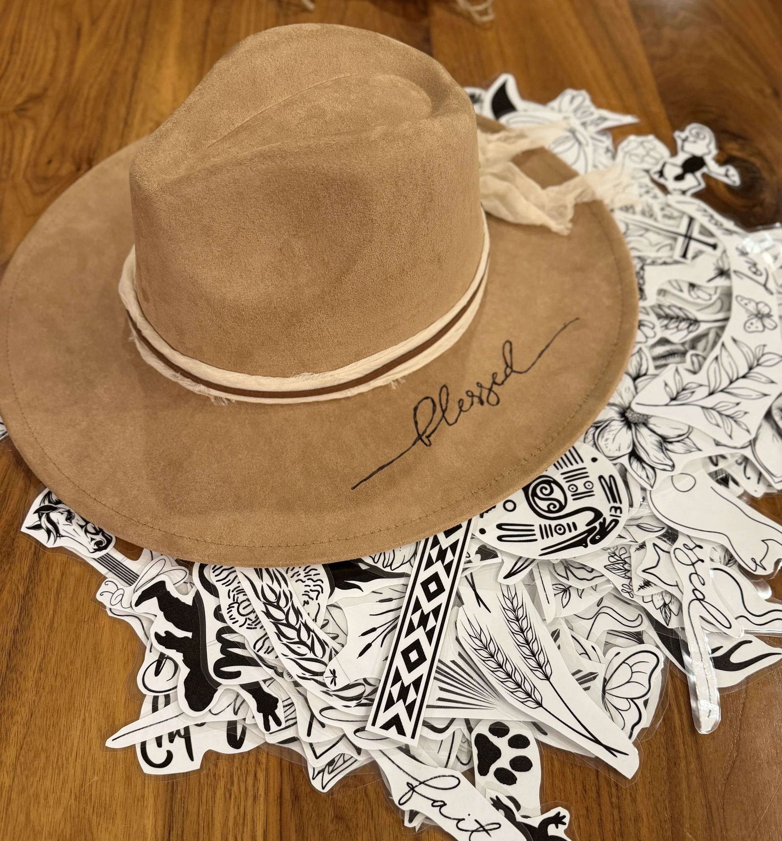 A stack of tan felt hats with a wide brim, placed on a surface covered with black-and-white sticker sheets featuring various illustrated images and designs.