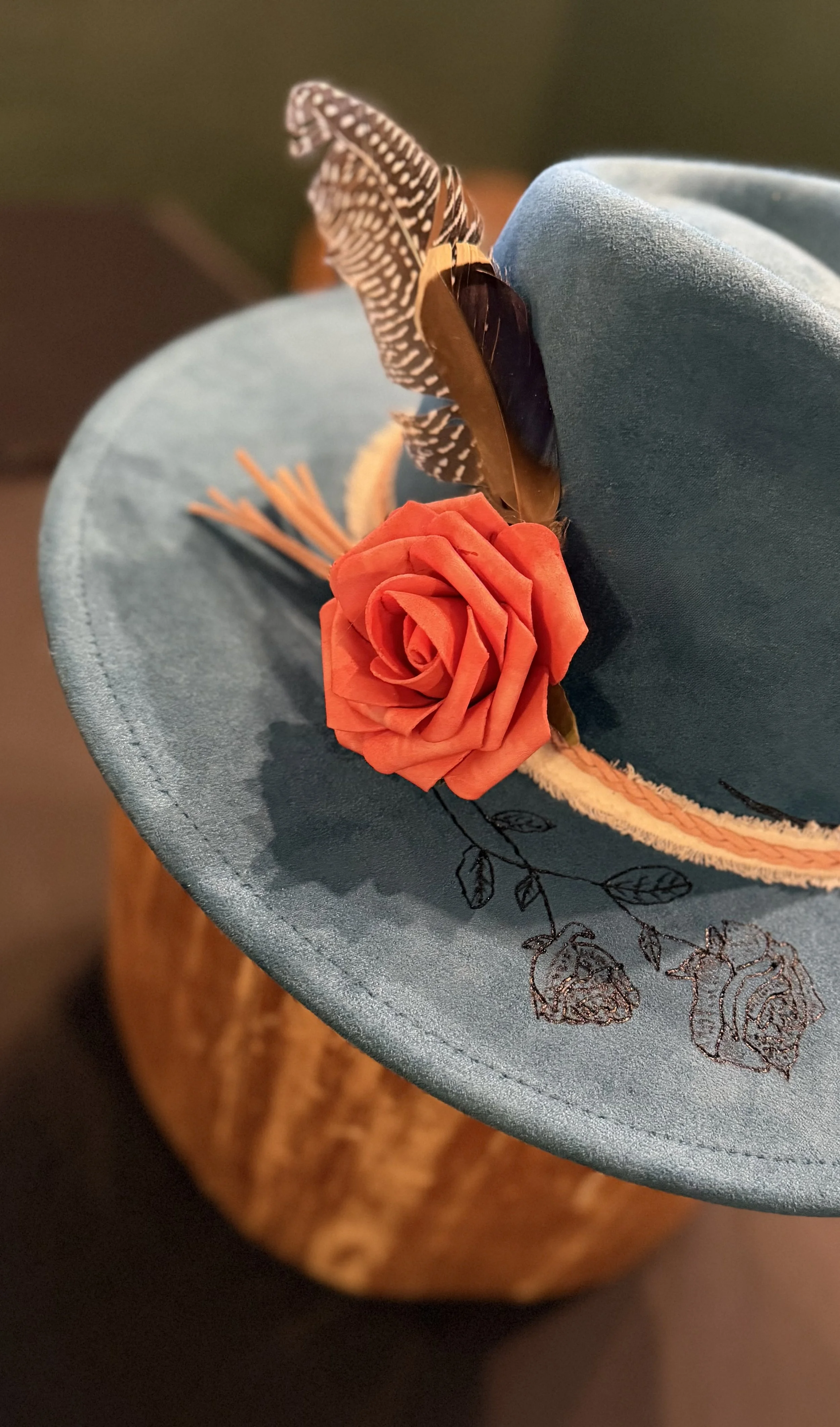 Close-up of a fashion hat with a blue brim, decorated with a red paper rose, a feather, and embroidery of roses and leaves.