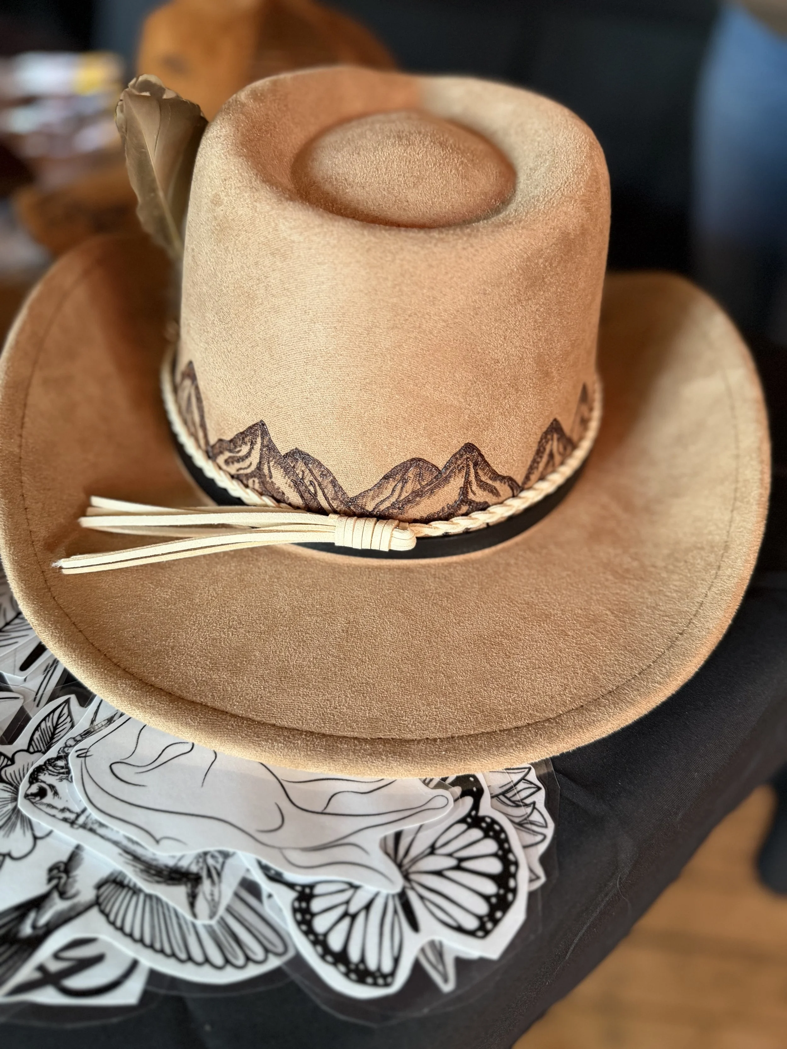 Close-up of a tan cowboy hat with a mountain landscape design around the band, resting on a surface with black and white floral paper underneath.