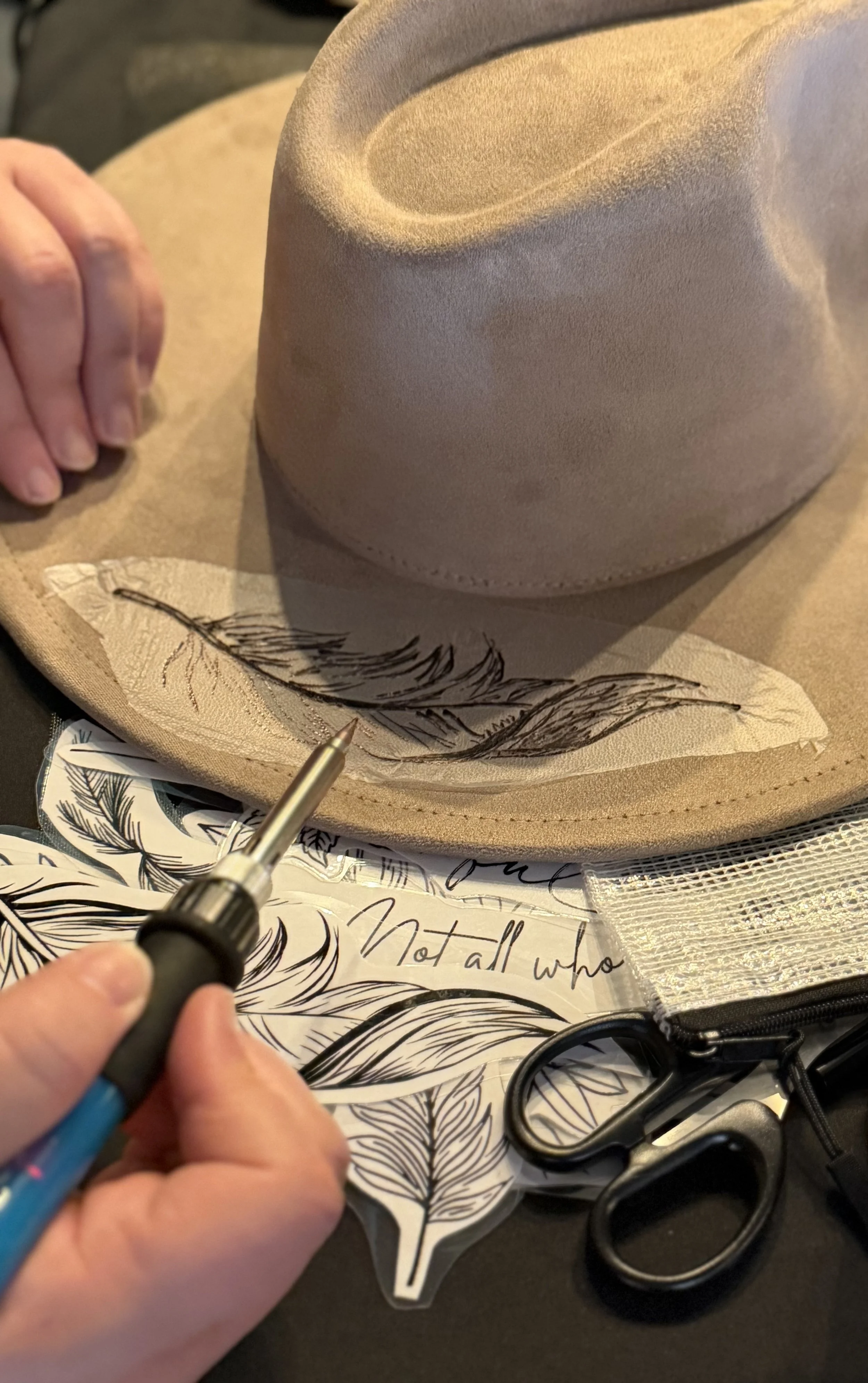 Person holding a marker with hand resting on a decorative cloth with a feather pattern, hat with a wide brim, and scissors around.