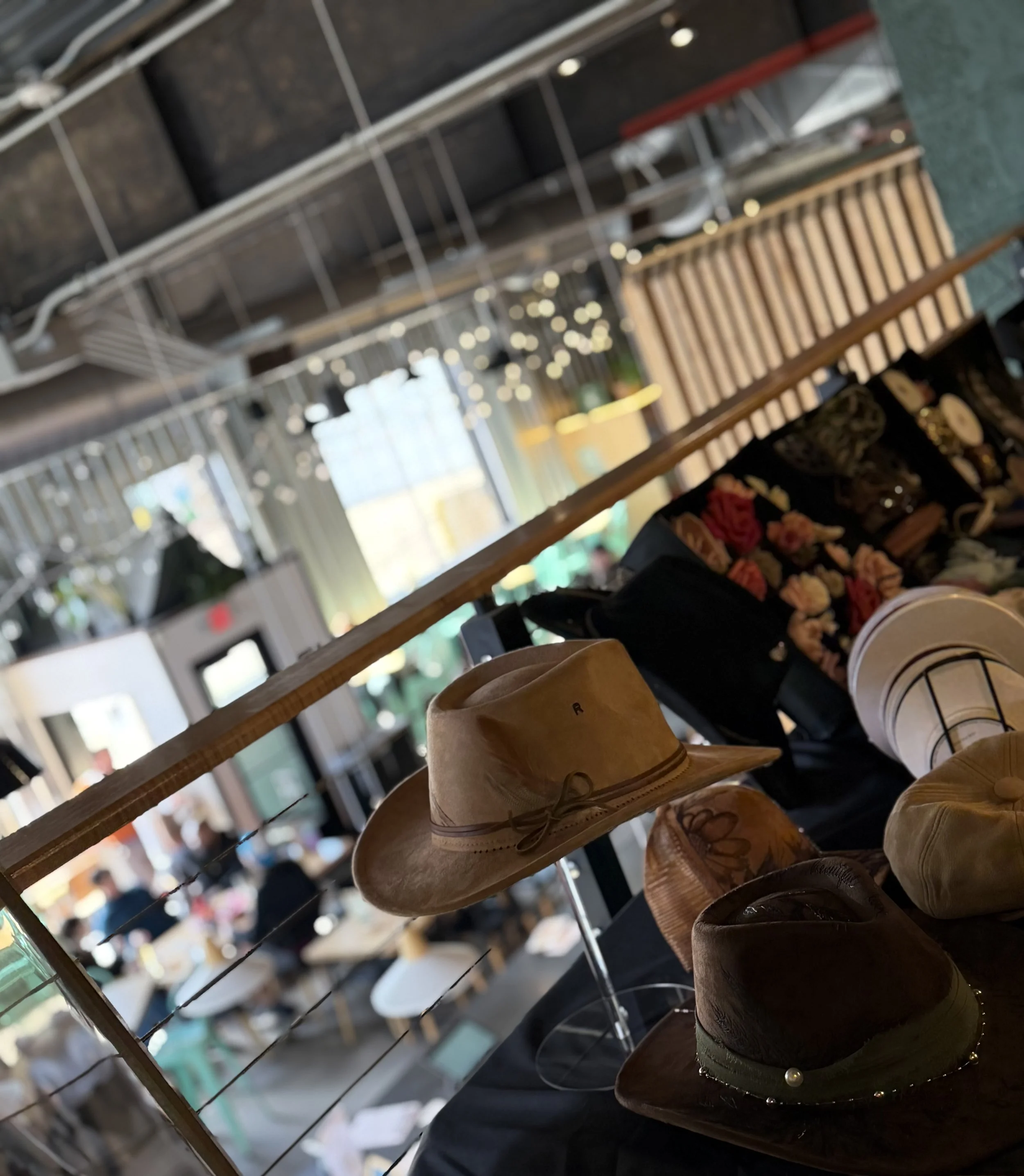 Display of various hats on a shelf in a retail store, including a tan fedora with a ribbon and pearl decoration, a dark brown hat, and a floral-print hat, with blurred background of store interior.