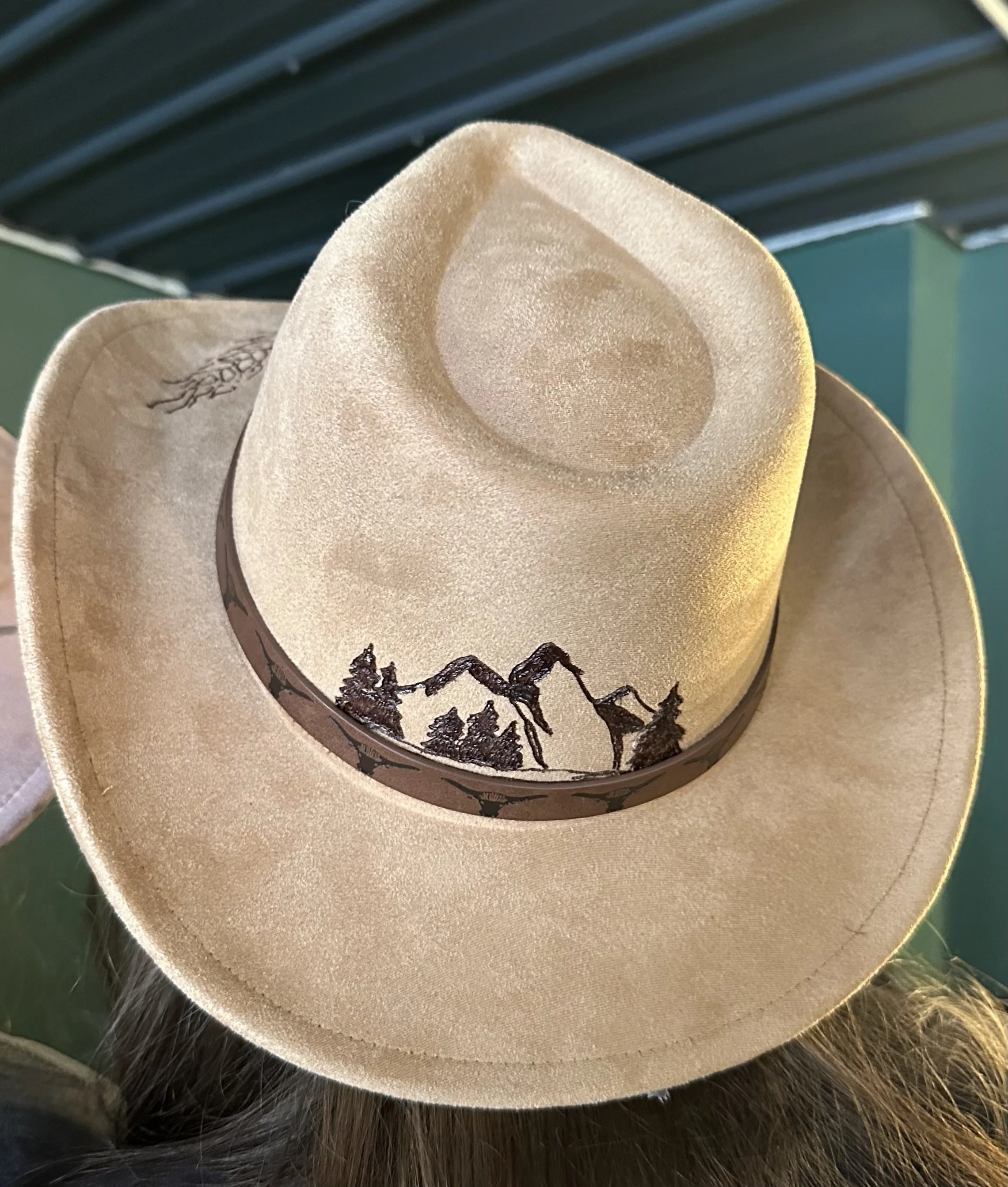 A beige cowboy hat with a drawn mountain scene and trees inside the crown, placed on a surface with dark and light brown fur surrounding it.