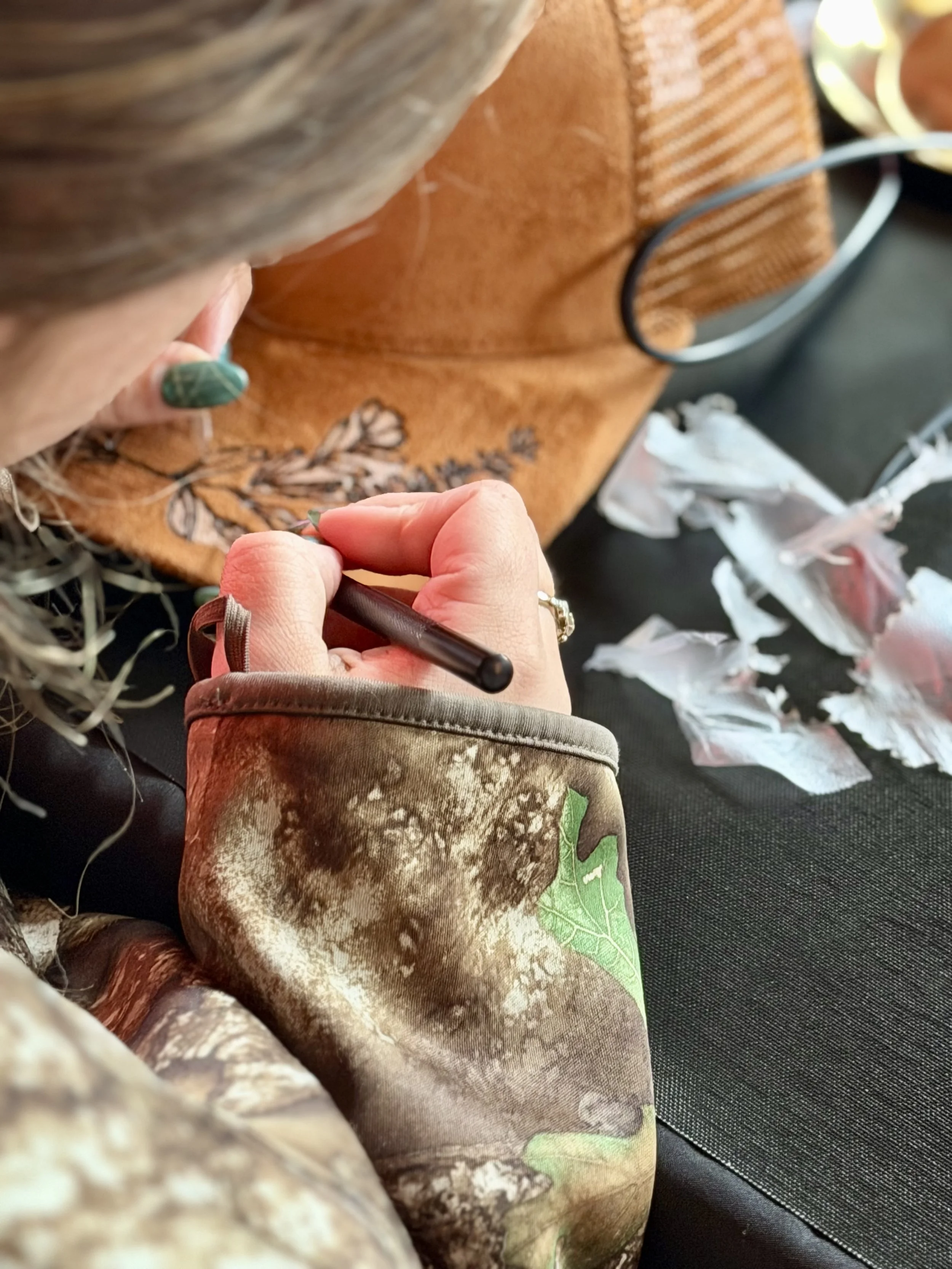 Close-up of a person sewing or drawing on fabric with a pen or stylus, with a focus on their hands and fabric. The person is wearing a beige or tan jacket with a pattern and has green painted nails. The fabric features an illustration of an animal, p