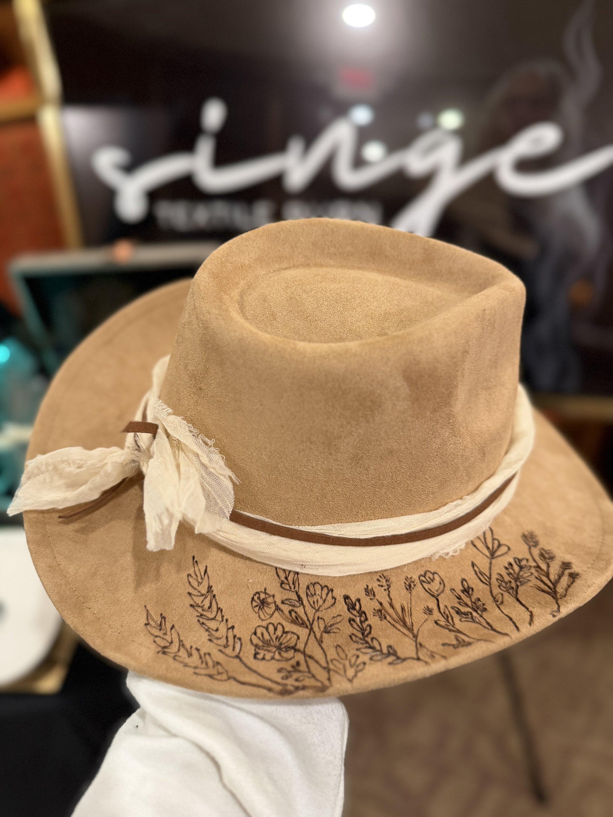 A beige felt hat with a wide brim decorated with black leaf and flower sketches, placed on a display stand with a white ribbon band and a small brown leather tag.