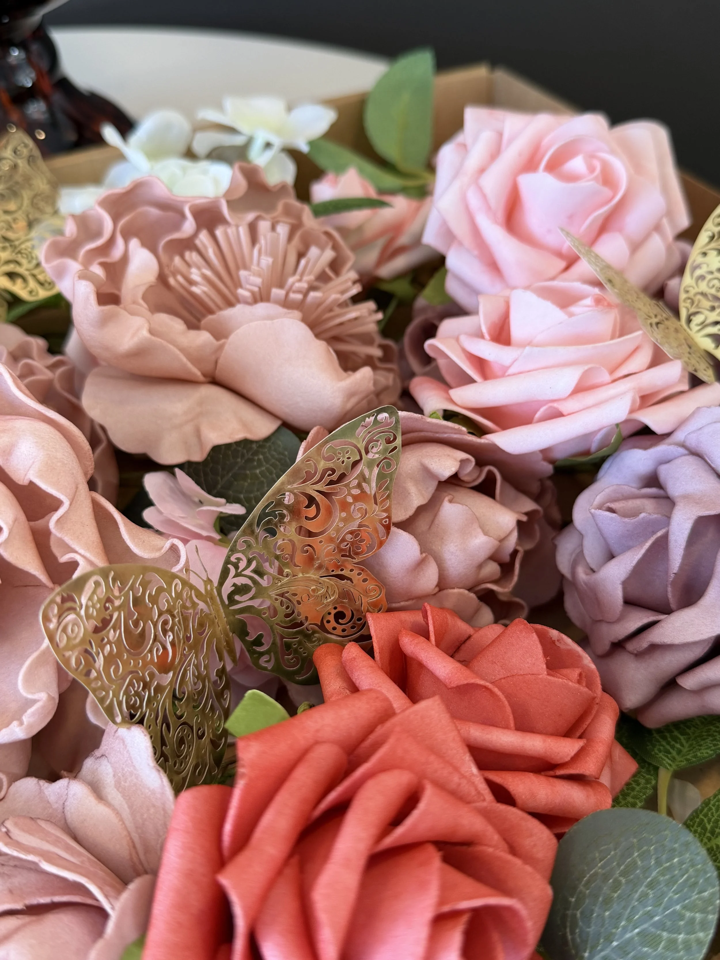 A bouquet of artificial pink, peach, and purple roses with green leaves, white flowers, gold decorative butterflies, and green foliage.
