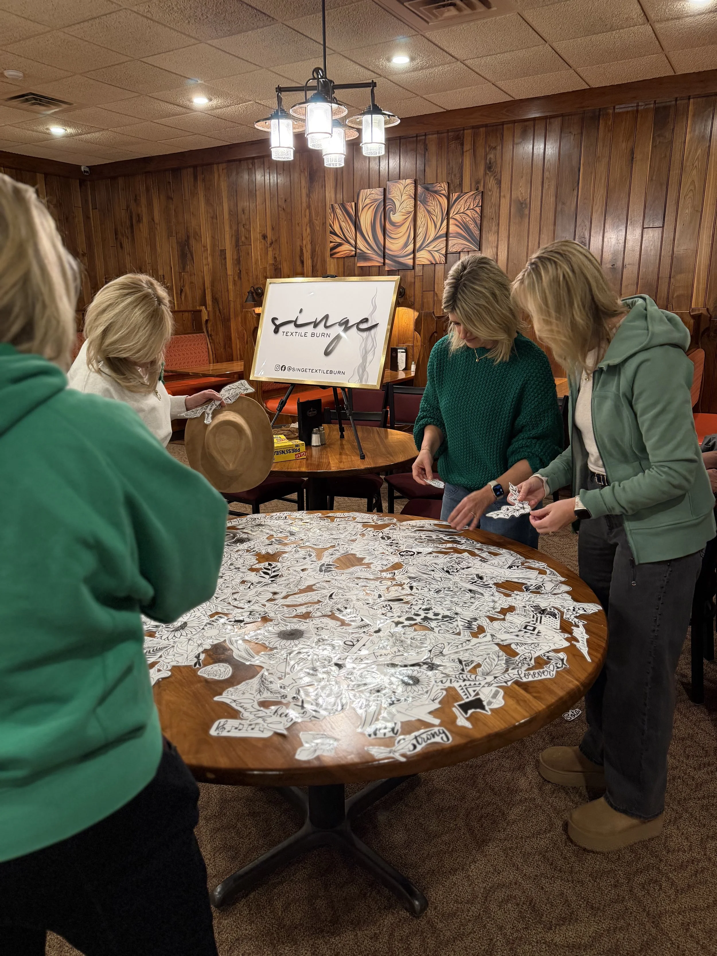 Several people gather around a wooden table covered with coloring pages and stickers in a room with wood-paneled walls and warm lighting. A sign on an easel reads 'Singe Textile Burn' with social media handles.