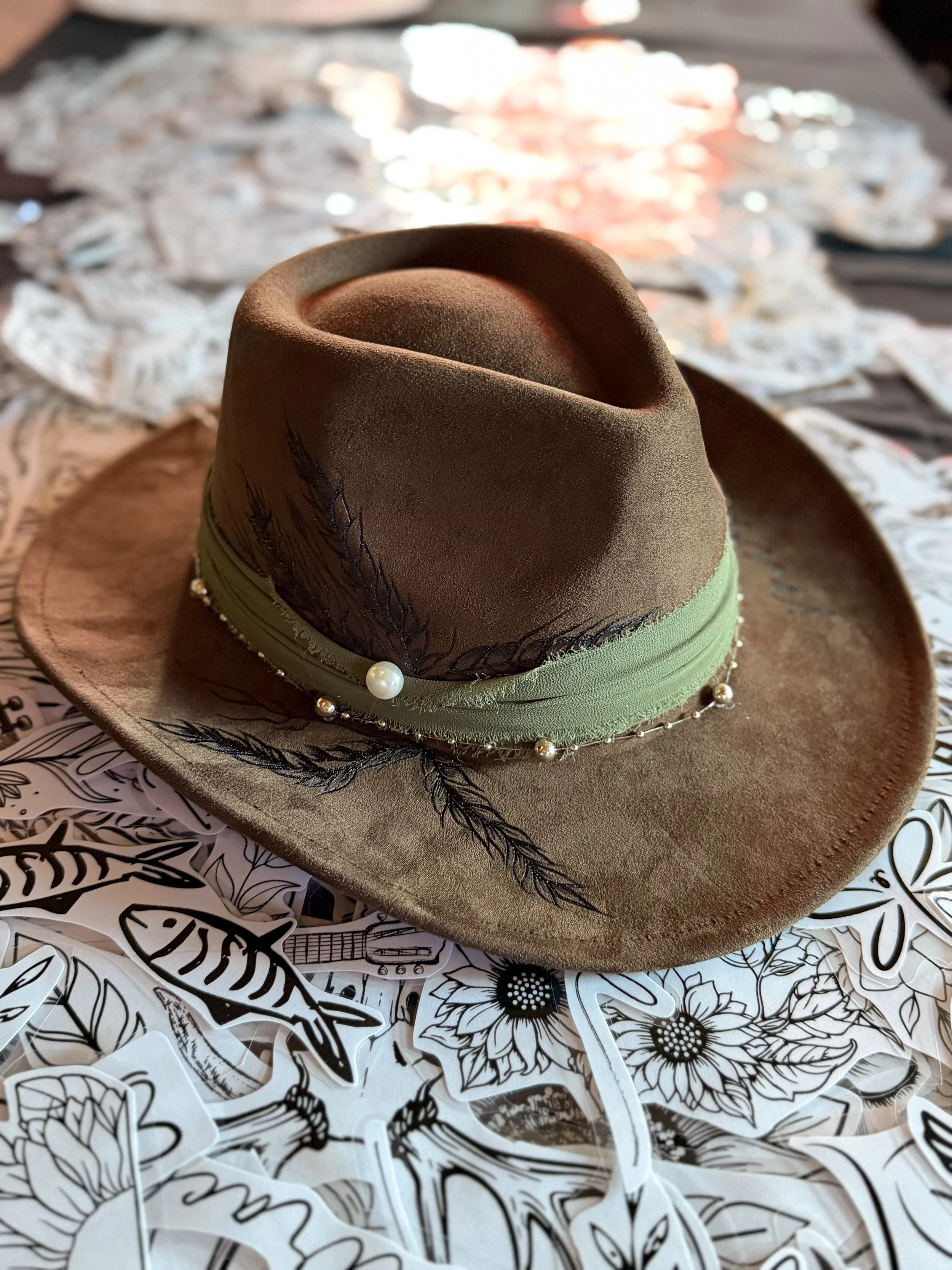 A brown felt cowboy hat decorated with black embroidered leaves and pearl accents, resting on a table covered with black and white floral patterned paper cutouts.