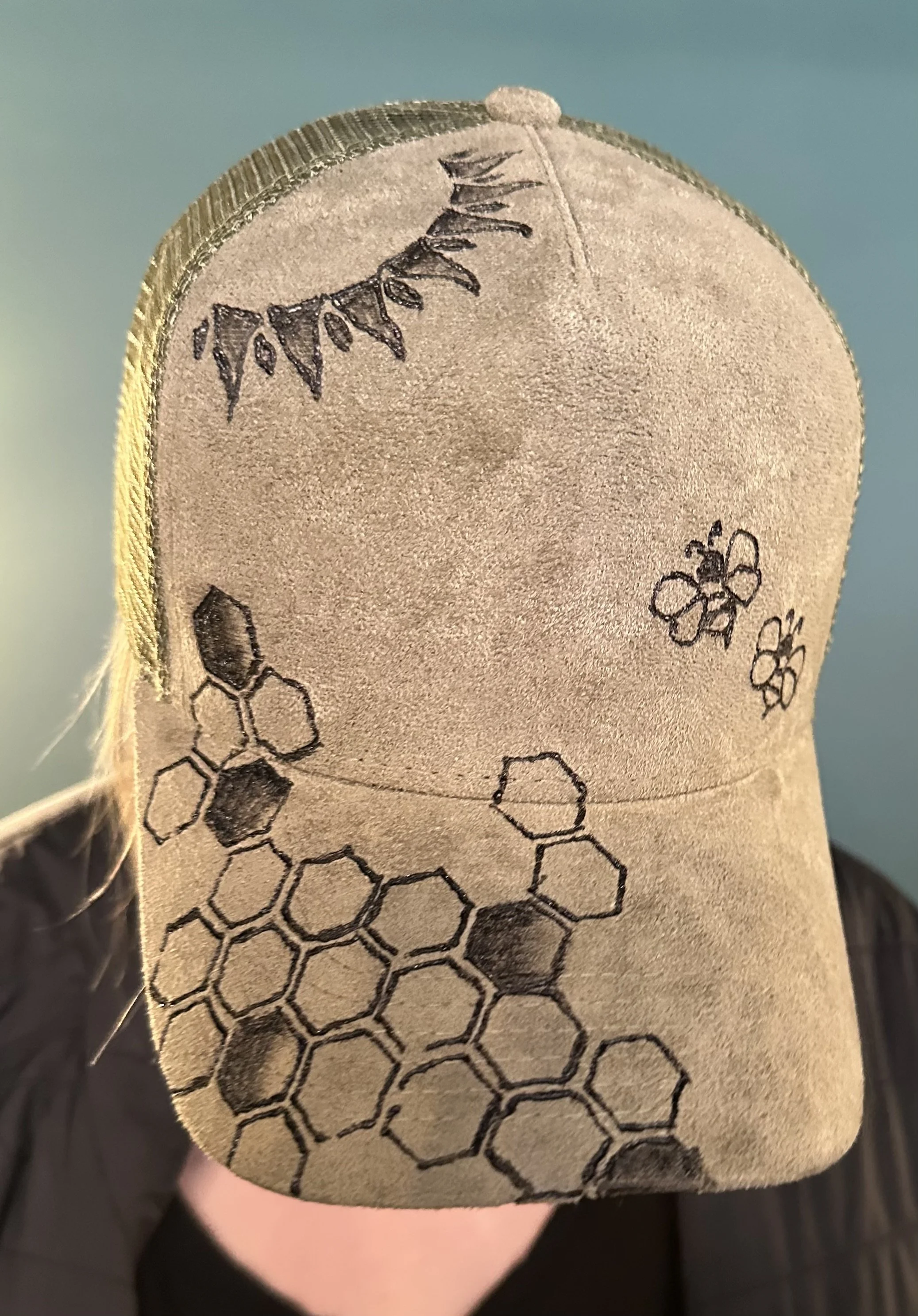 A tan baseball cap with black printed illustrations of honeycomb patterns, bees, and a branch with leaves and flowers.
