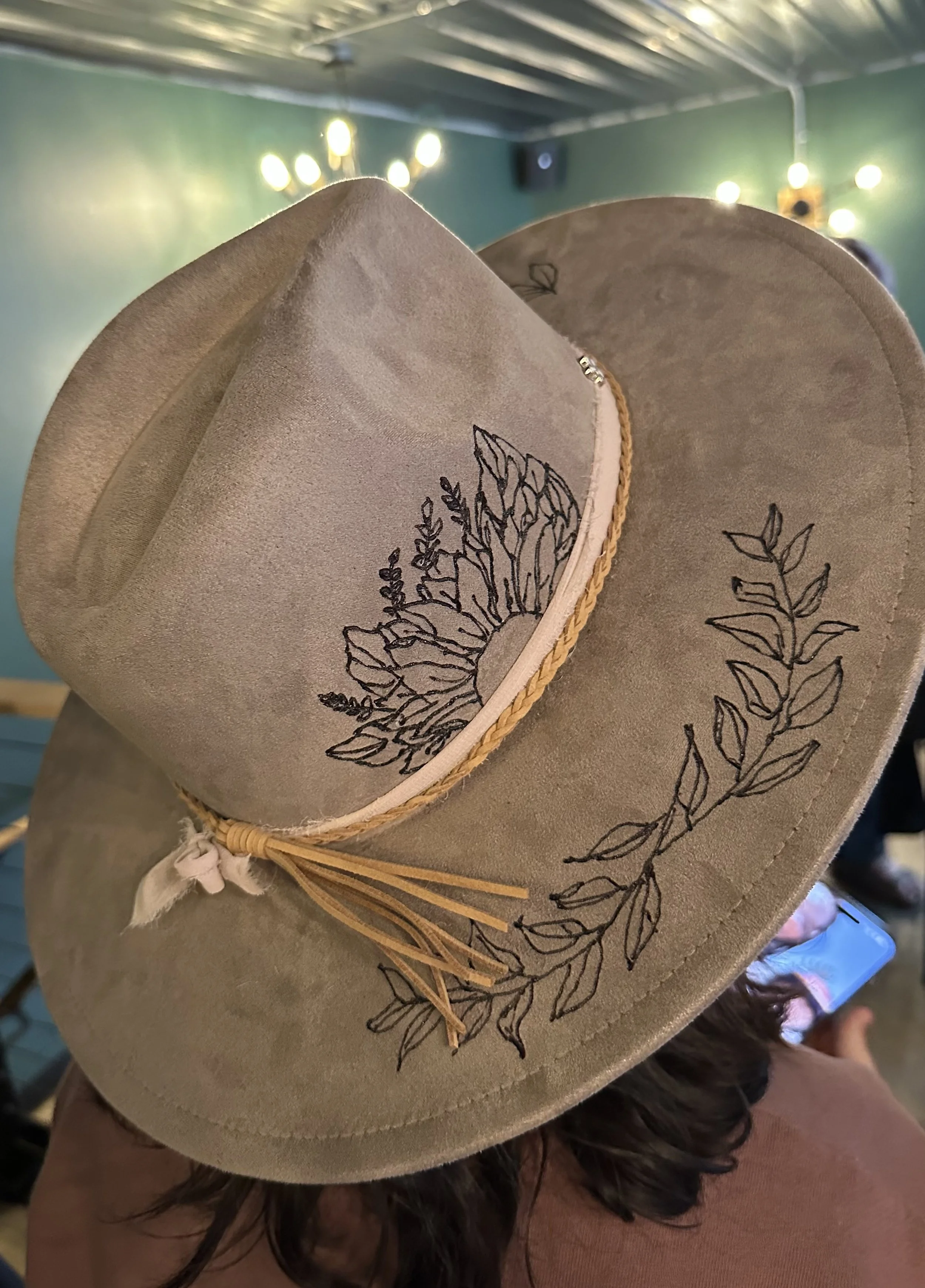 A person wearing a beige cowboy hat with black embroidered leaf patterns, a braided band, and a ribbon tied at the back, in an indoor setting with green walls and string lights in the background.