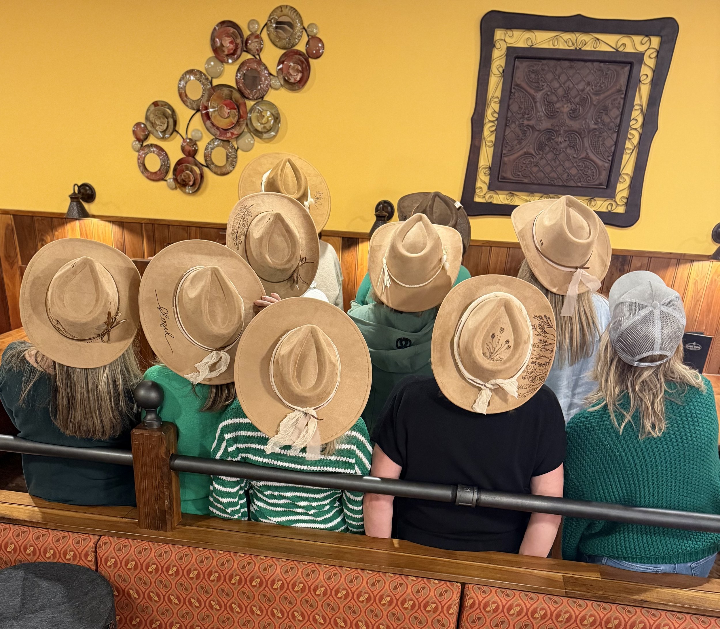 A group of people wearing tan cowboy hats with decorative bands, gathered in a restaurant with a yellow wall and wooden decor. Some individuals are seen from behind, and one person has a green and white striped shirt. There is a wall decoration on th