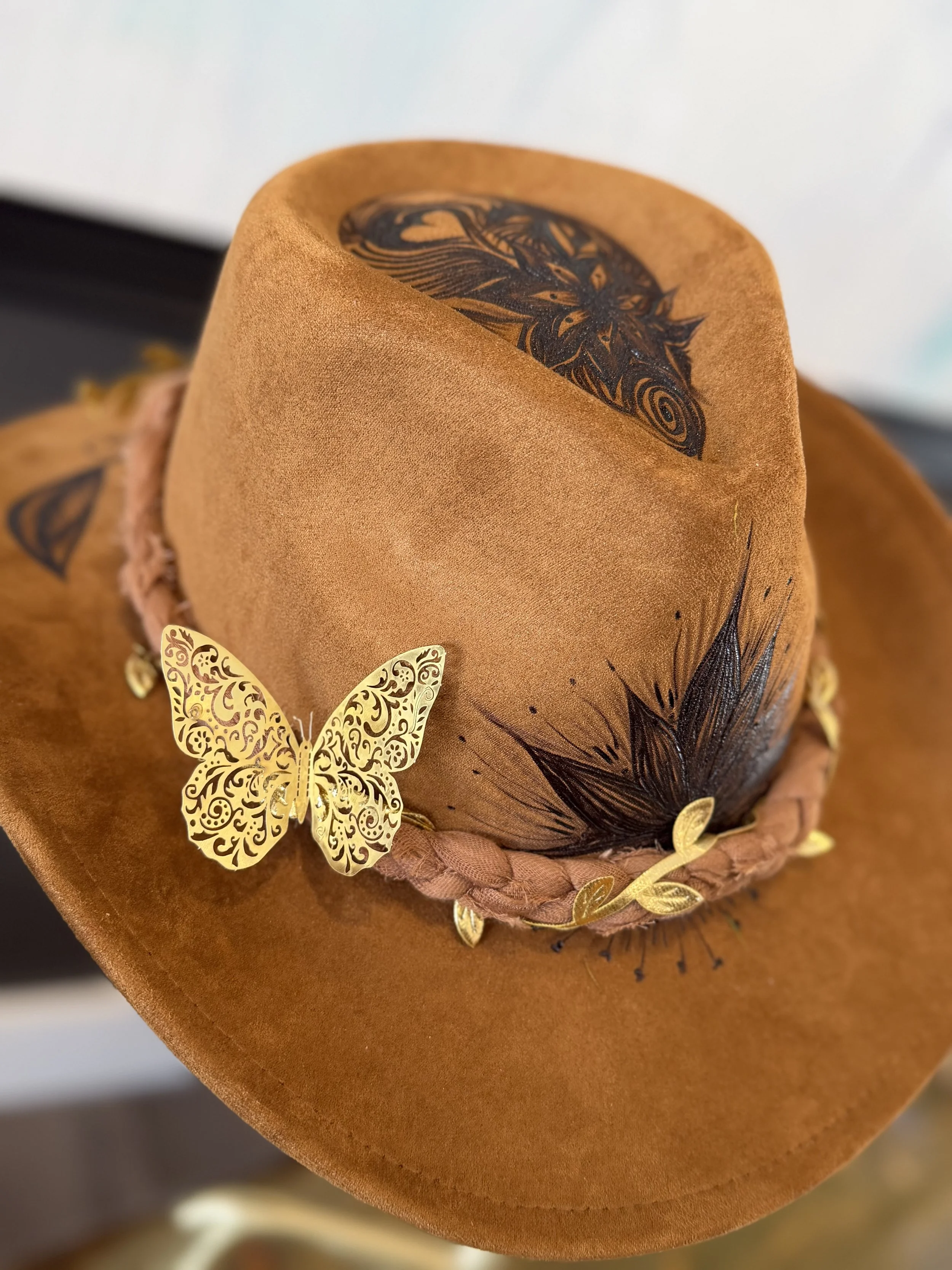 A close-up view of a brown felt hat decorated with black feathered accents, a gold butterfly, and gold leaf-shaped embellishments.