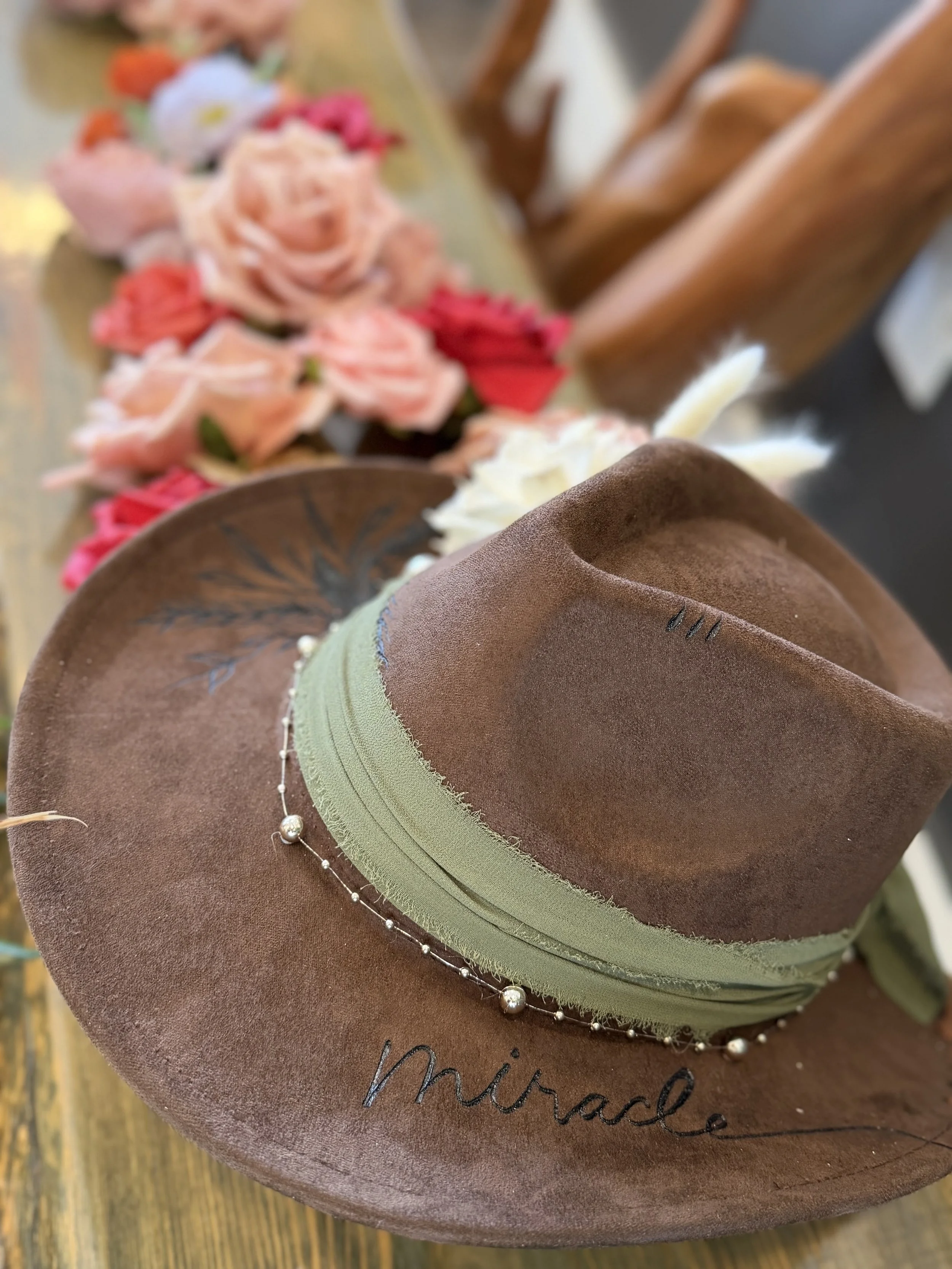 A brown cowboy hat with a green band and a pearl necklace resting on it, placed on a wooden surface. The hat has the word 'Miracle' embroidered on it.