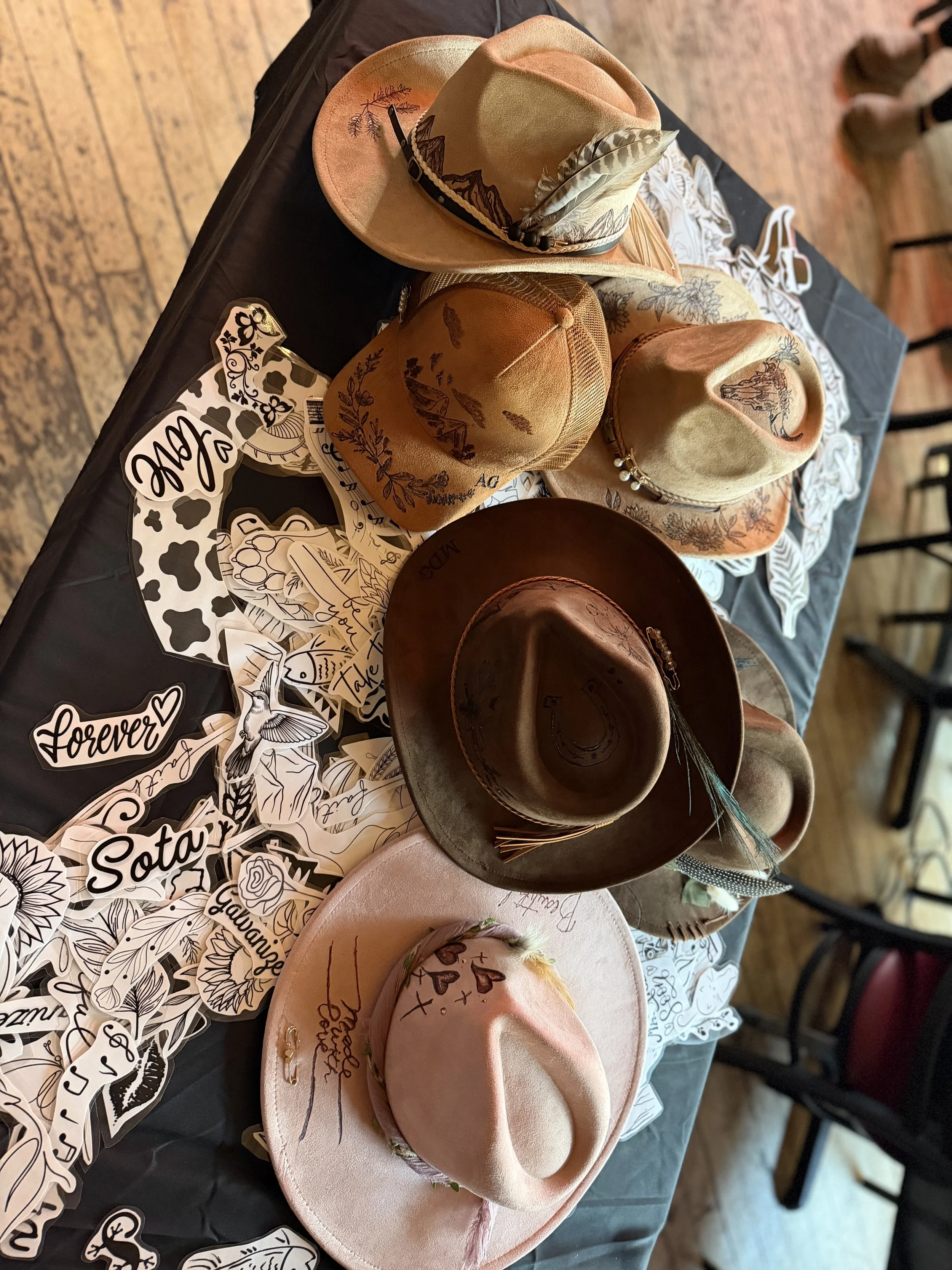 Several cowboy hats displayed on a table, some decorated with feathers, embroidery, and drawings, alongside paper stickers with various designs and words.