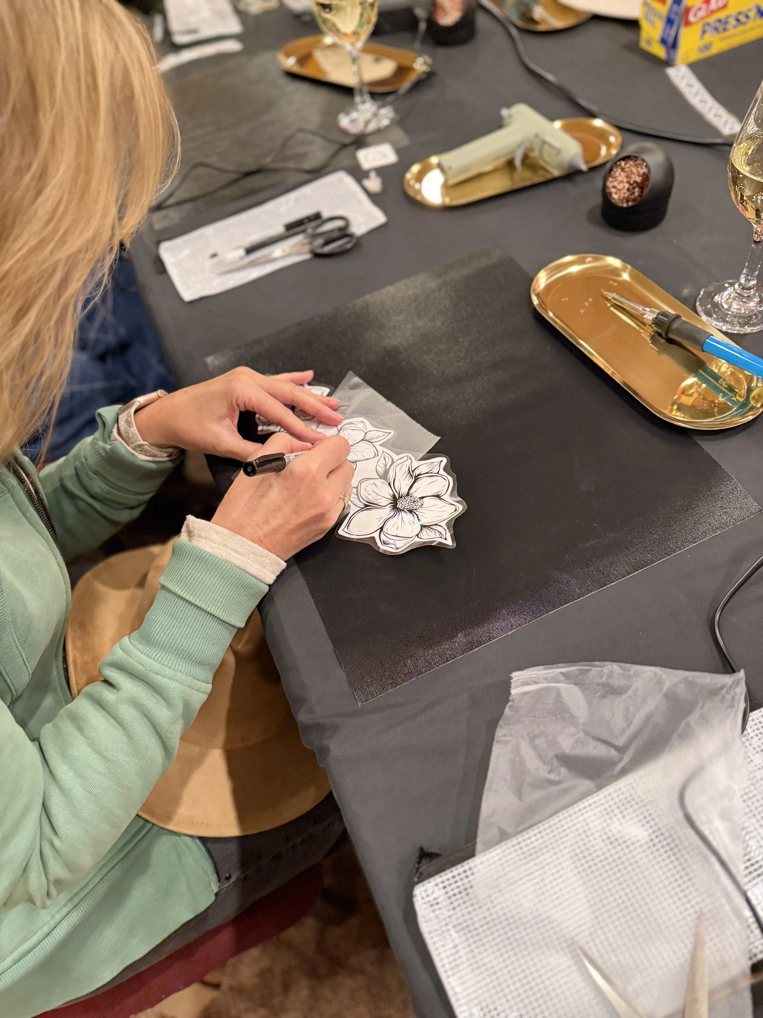 A person with blonde hair and a green jacket is coloring a flower pattern on a piece of paper with a black marker. The table has various art supplies, including a gold tray, a glass of white wine, scissors, and a box of animal crackers.