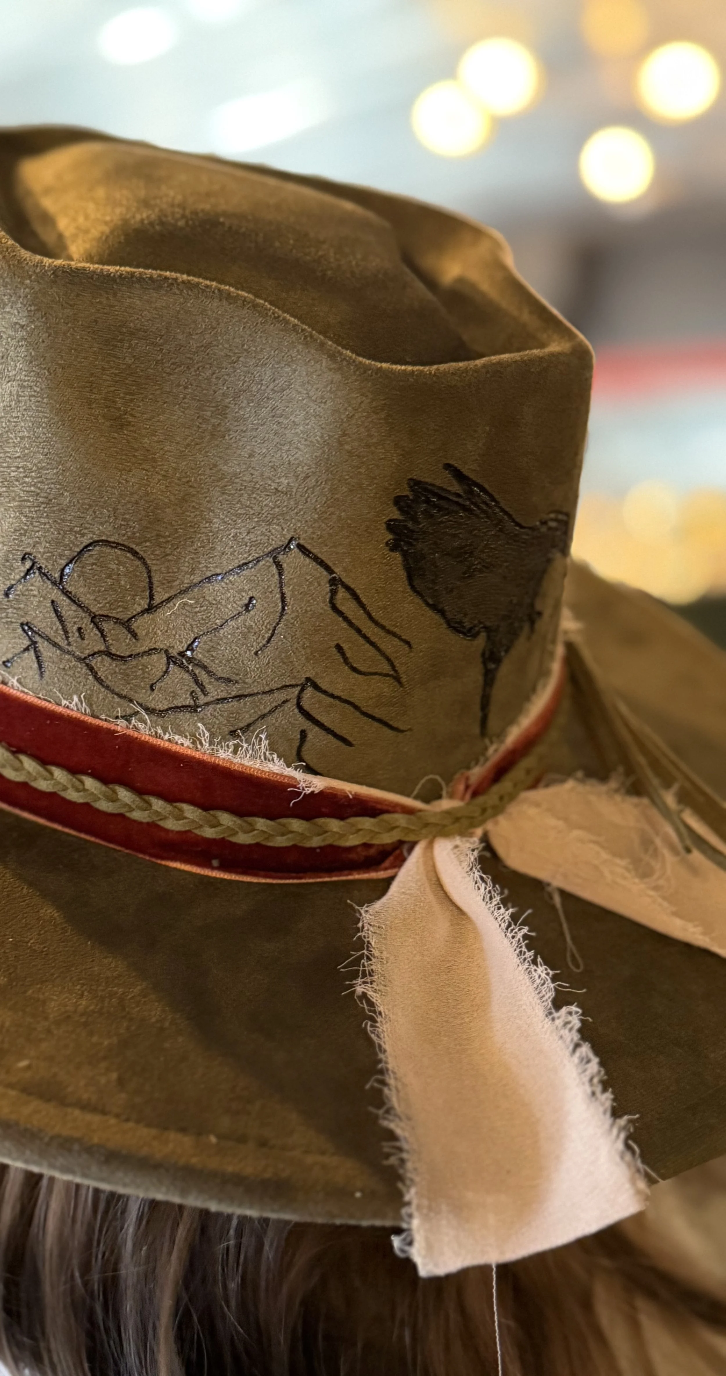 Close-up of a cowboy hat with embroidered design, decorative band, and feathers, with a blurred background of lights.