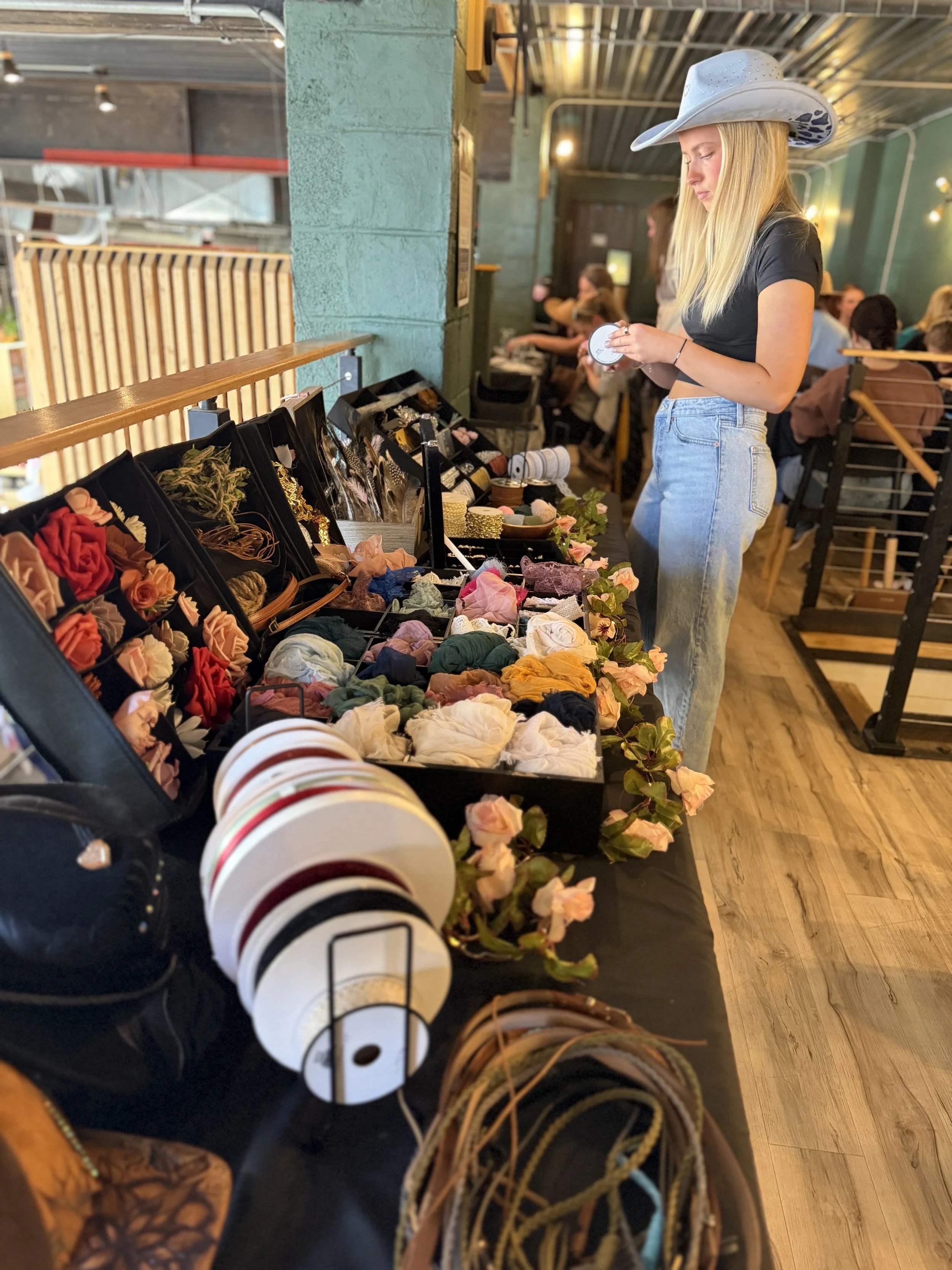 A woman wearing a white cowboy hat and black crop top shopping at a store with accessories, including hair scrunchies, headbands, jewelry, and hats, displayed on a table.