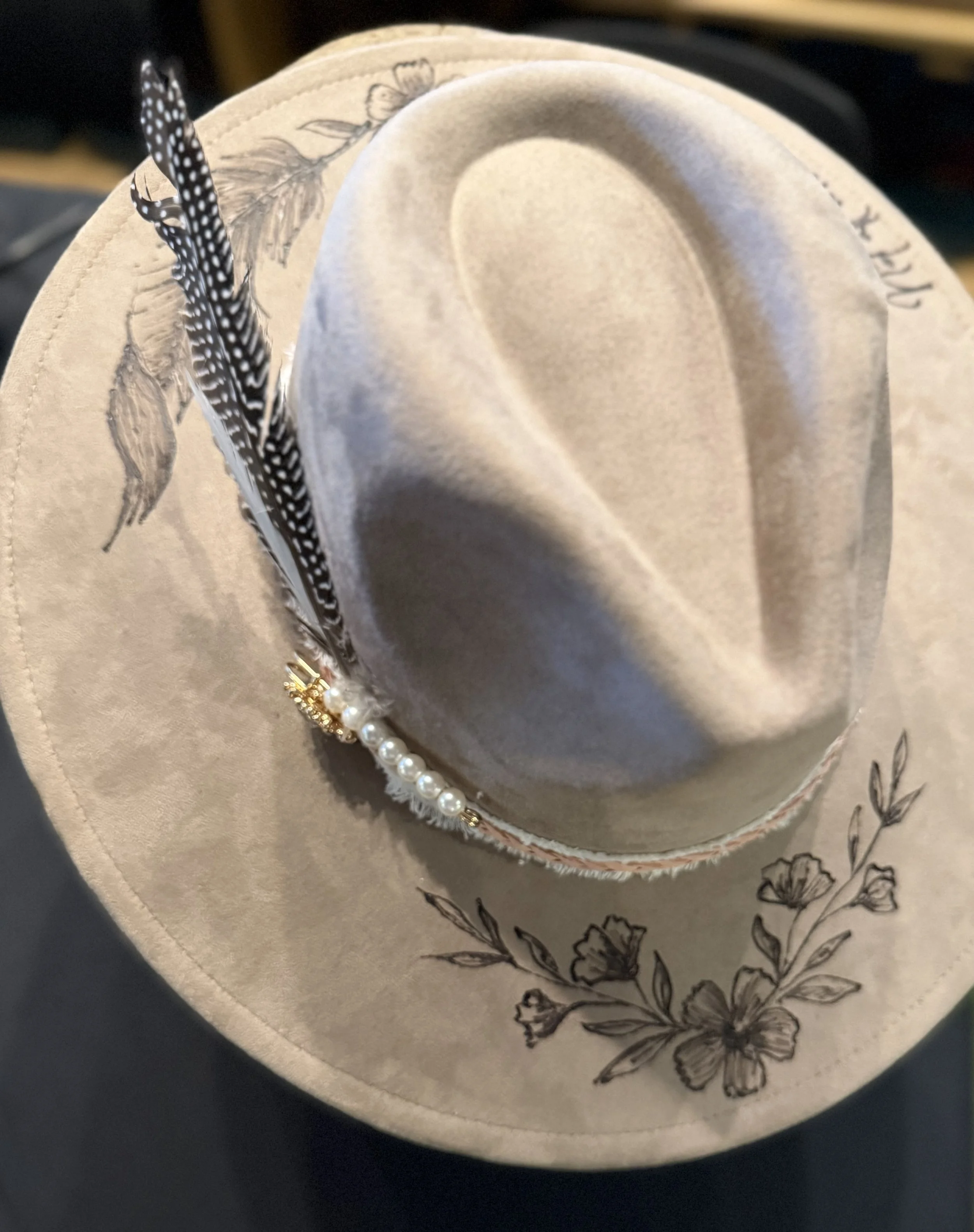 A beige hat with black floral embroidery on the brim, decorated with a string of white pearls and small gold accents, resting inside a hat box.