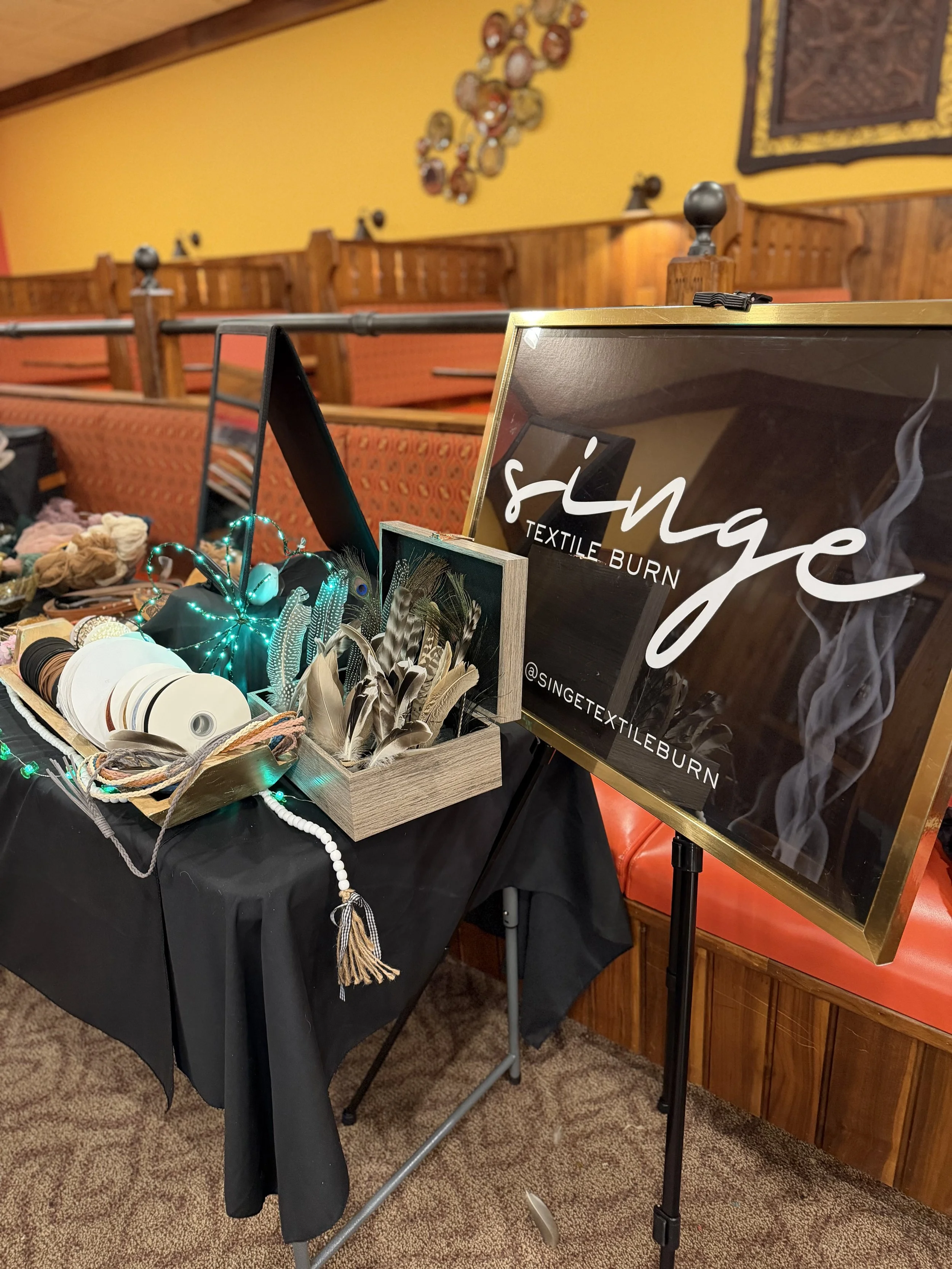 Indoor display table with textiles, feathers, and decorative items, next to a sign reading '@SINGETEXTILEBURN'.