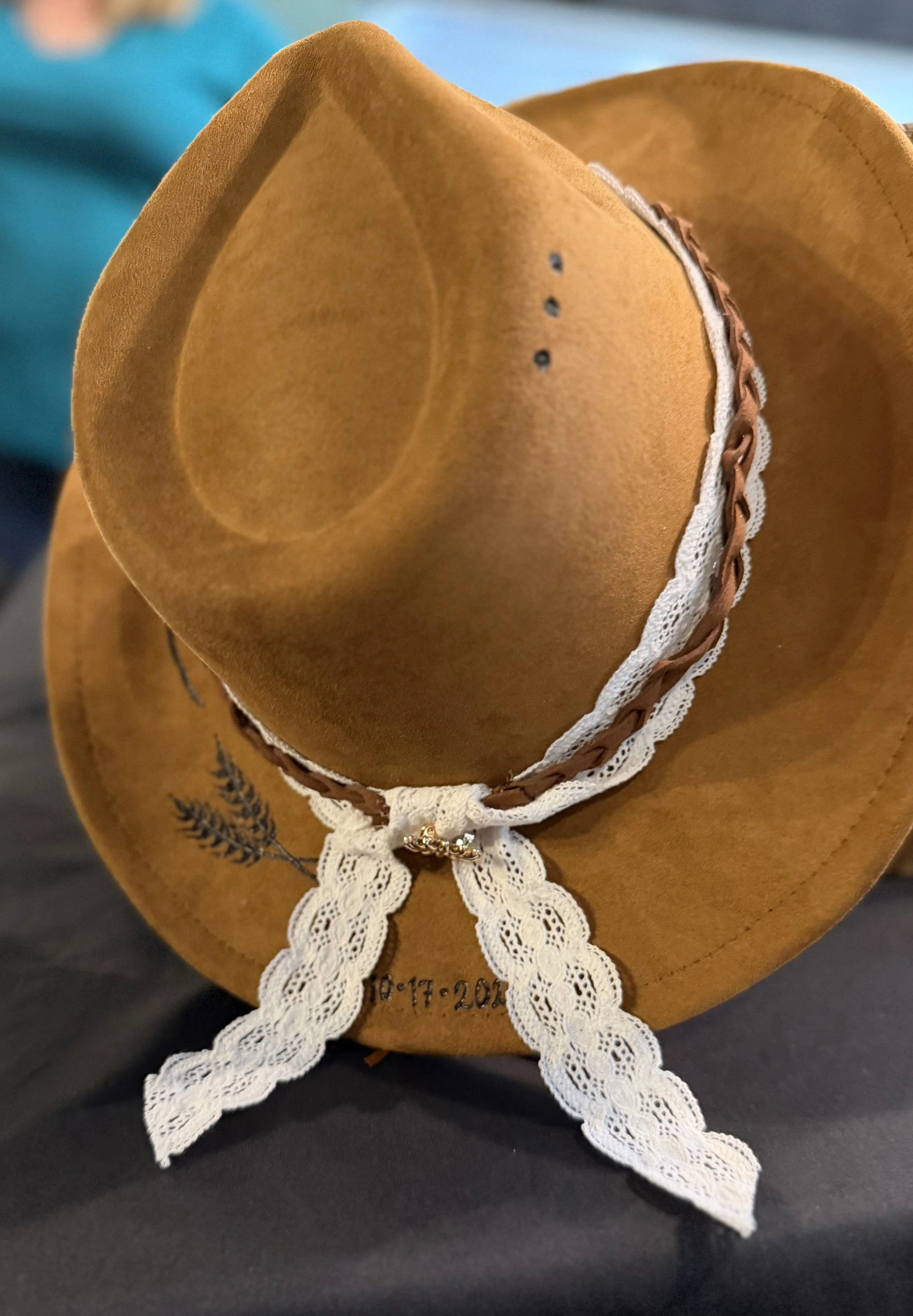 A brown cowboy hat with white lace trim and embroidered black plant design, resting on a dark surface.