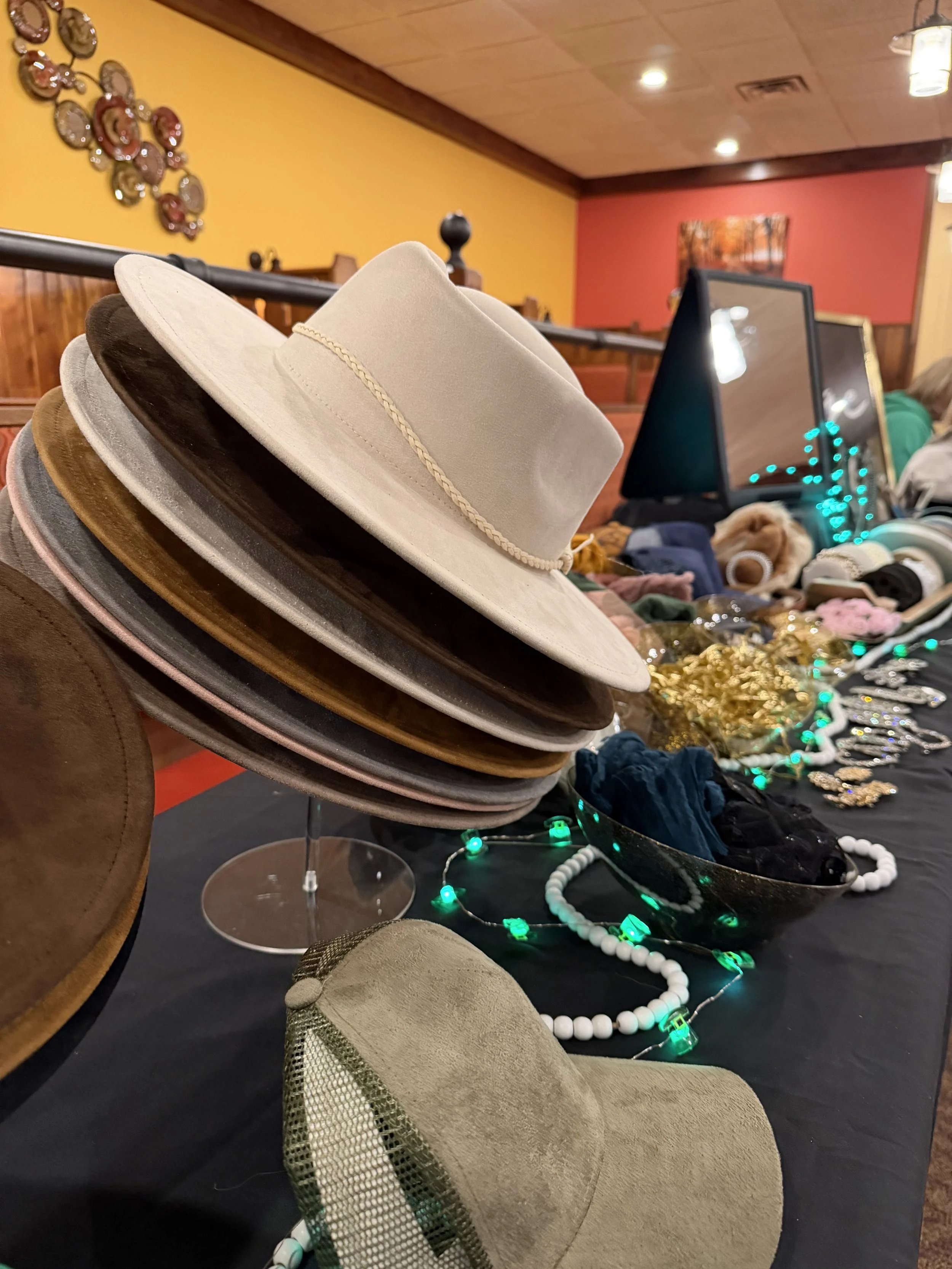 Stacked hats on display table at an indoor market, with necklaces, jewelry, and decorative items on the table, and colorful walls in the background.