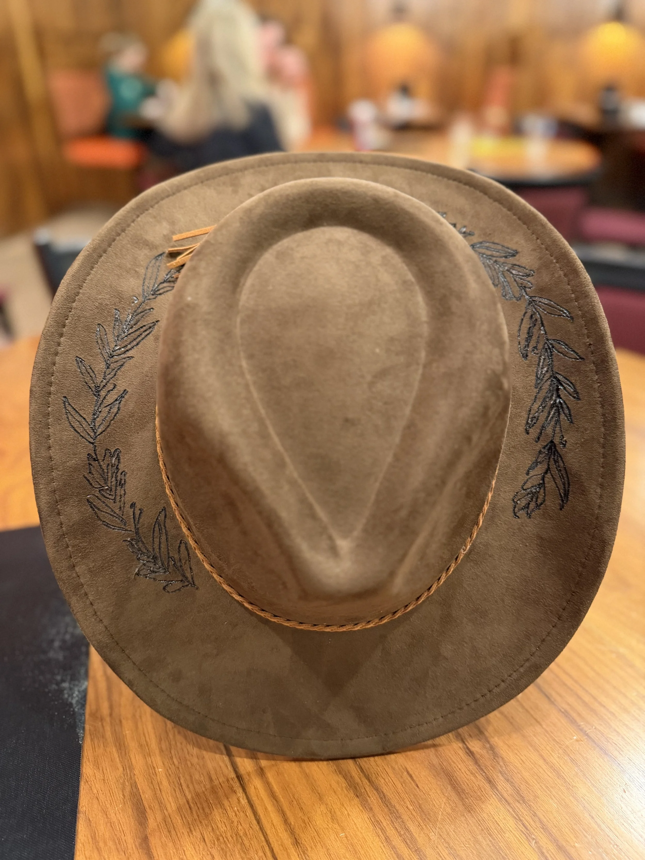 A brown cowboy hat with embroidered leaf designs around the brim, placed on a wooden table in a restaurant setting.