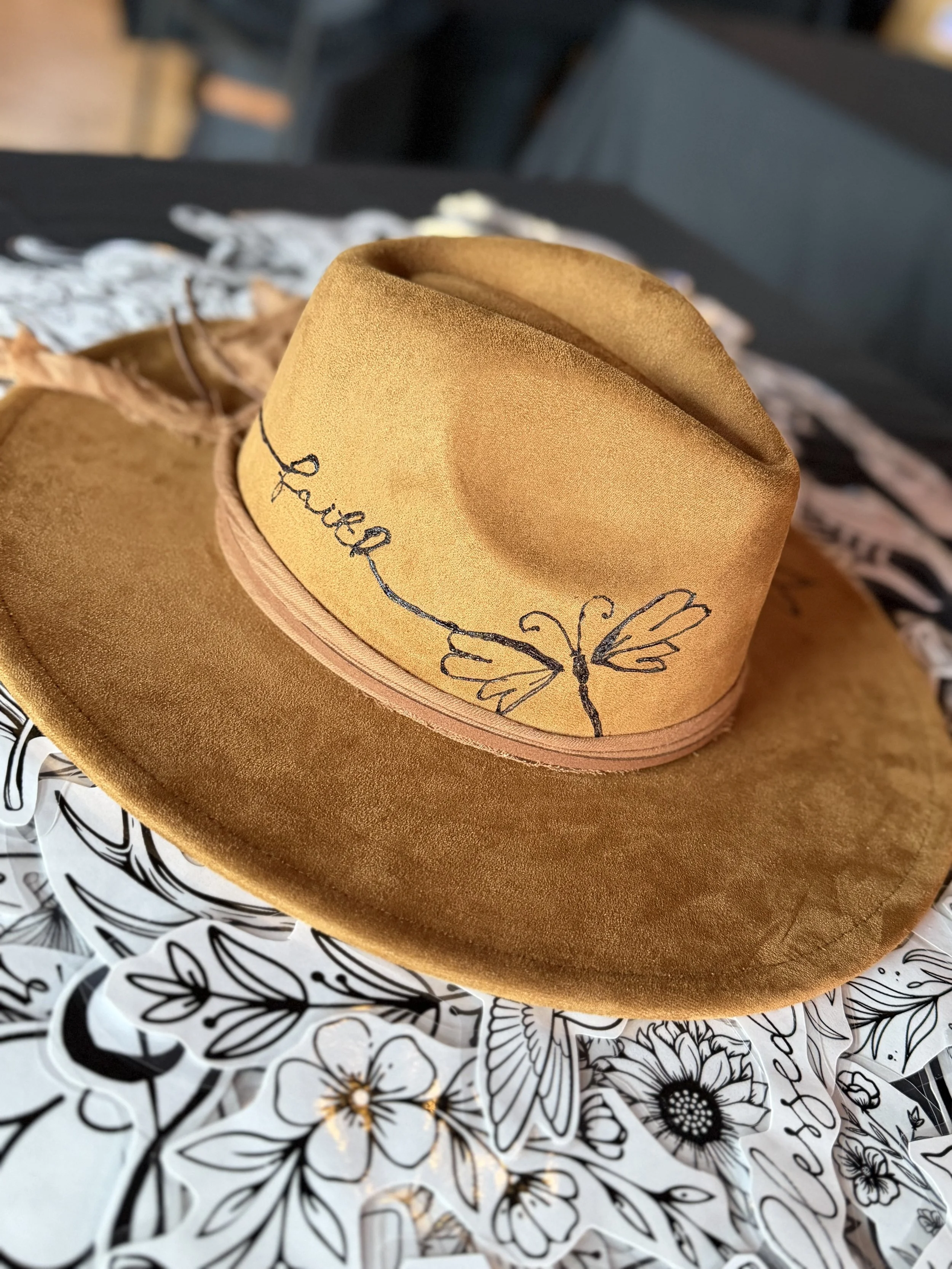 A brown felt hat with a embroidered butterfly and the words 'proud' and 'you' on the band, resting on a black and white floral patterned surface.