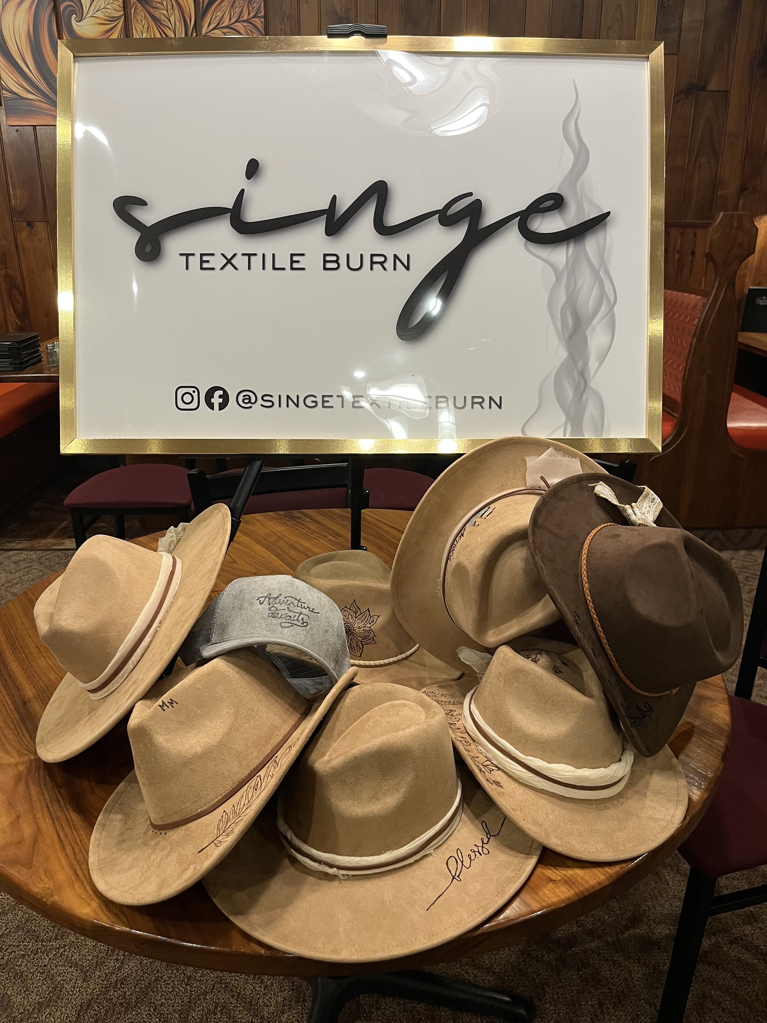 Various cowboy hats displayed on a wooden table in front of a sign that reads 'singe TEXTILE BURN' with social media icons and '@SINGETEXILEBURN' at the bottom.
