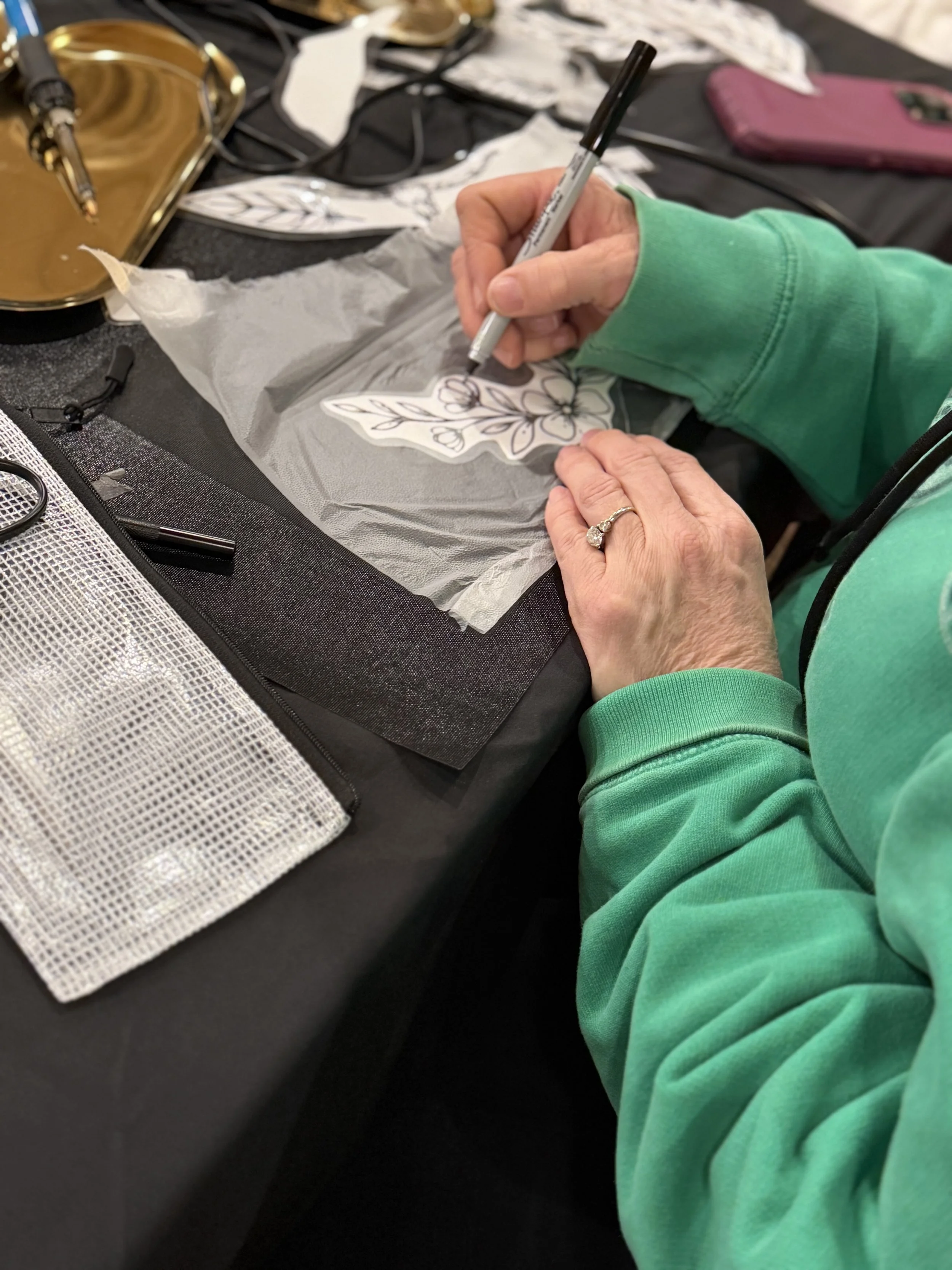 A person wearing a green long-sleeve shirt is drawing a floral design on a paper cutout with a black marker. The workspace includes a gold pen case, a pink phone, and various art supplies on a black table.