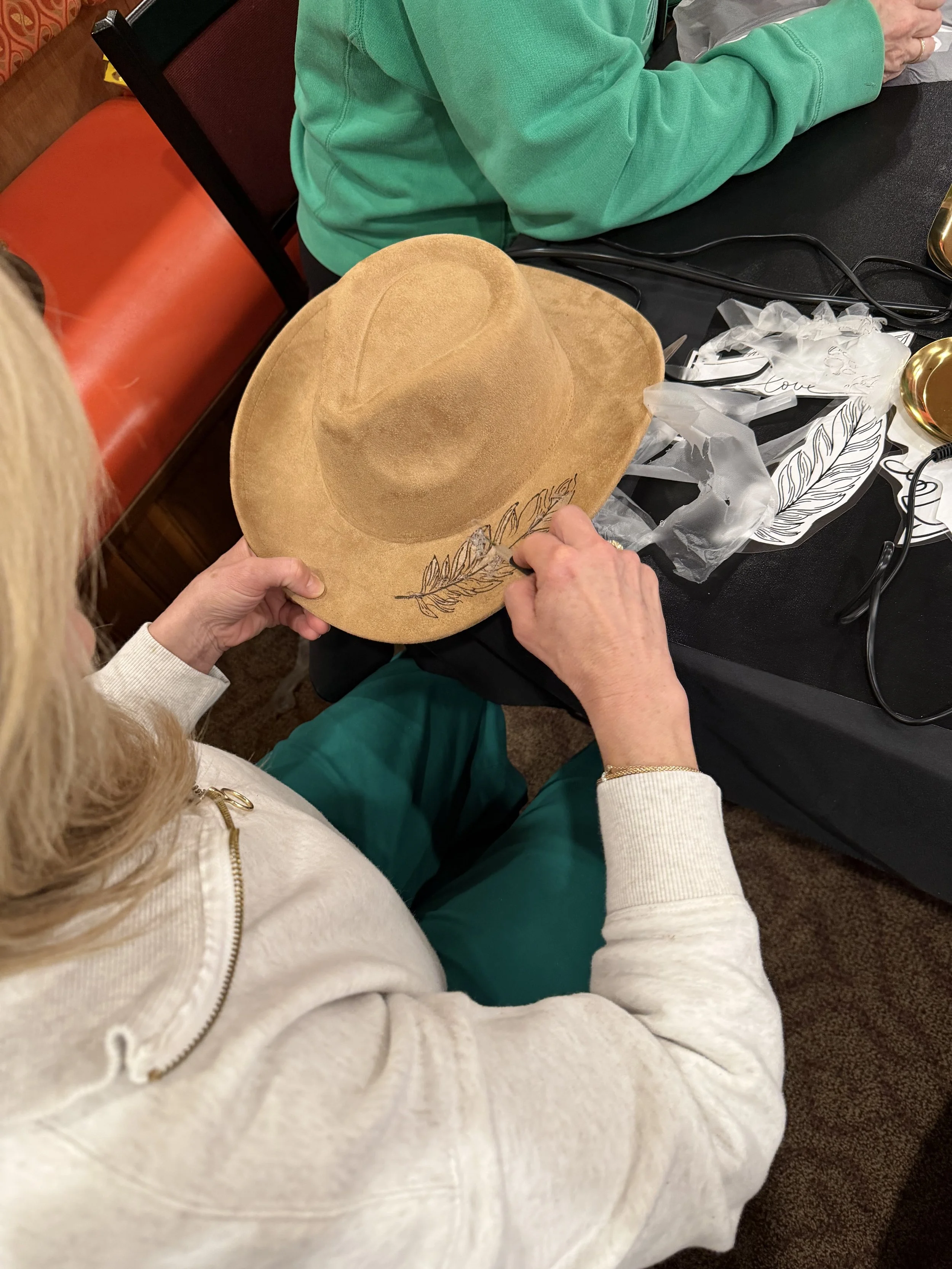 A person wearing a white hoodie sitting at a table with a tan fedora hat that has a feather print design on it. The person is holding the hat and drawing or writing on it with a black marker. There are other people seated at the table, one wearing a 
