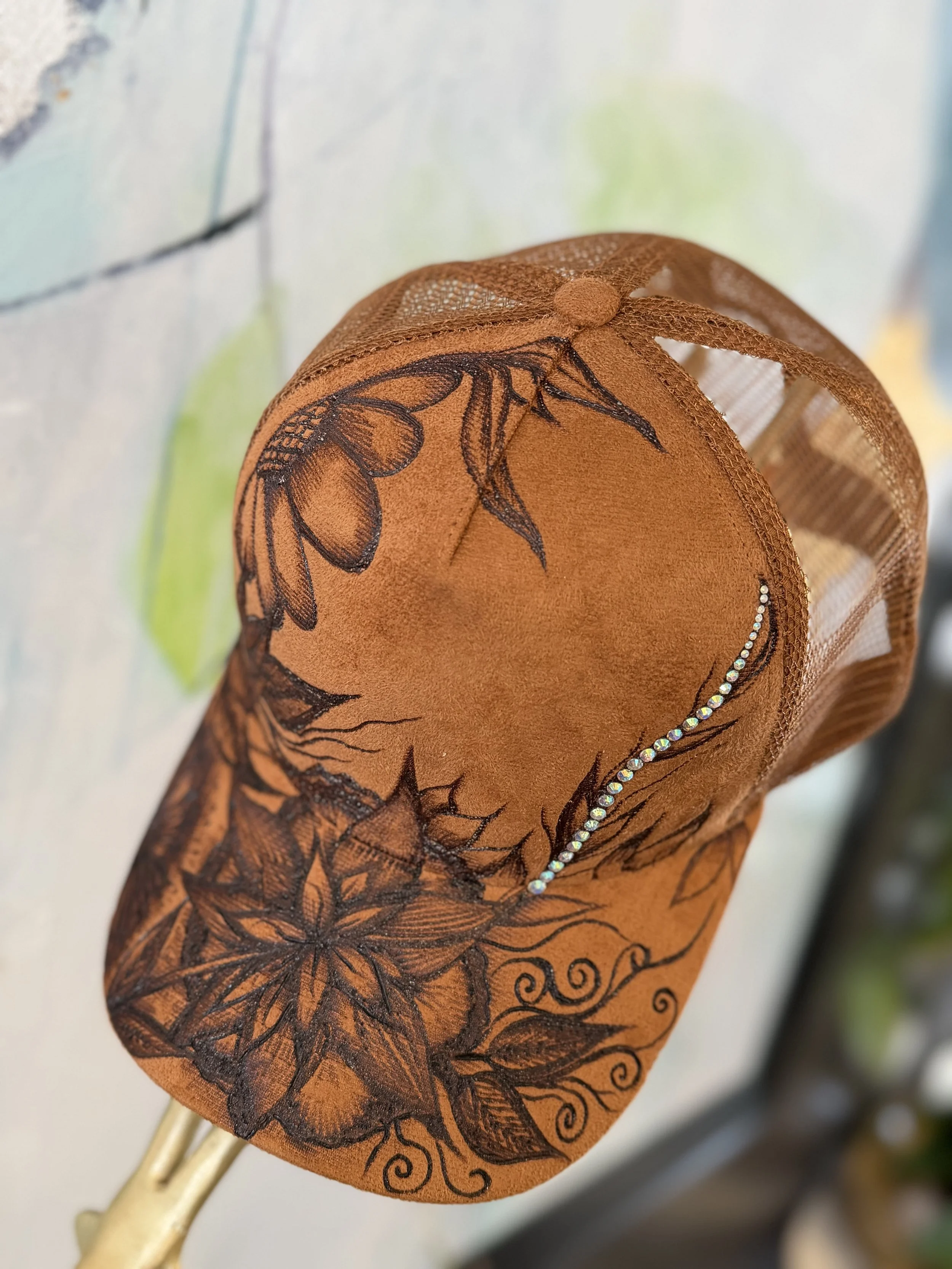 A decorative brown ornament with black floral and leaf designs, embellished with small rhinestones along one edge, hanging on a white wall.