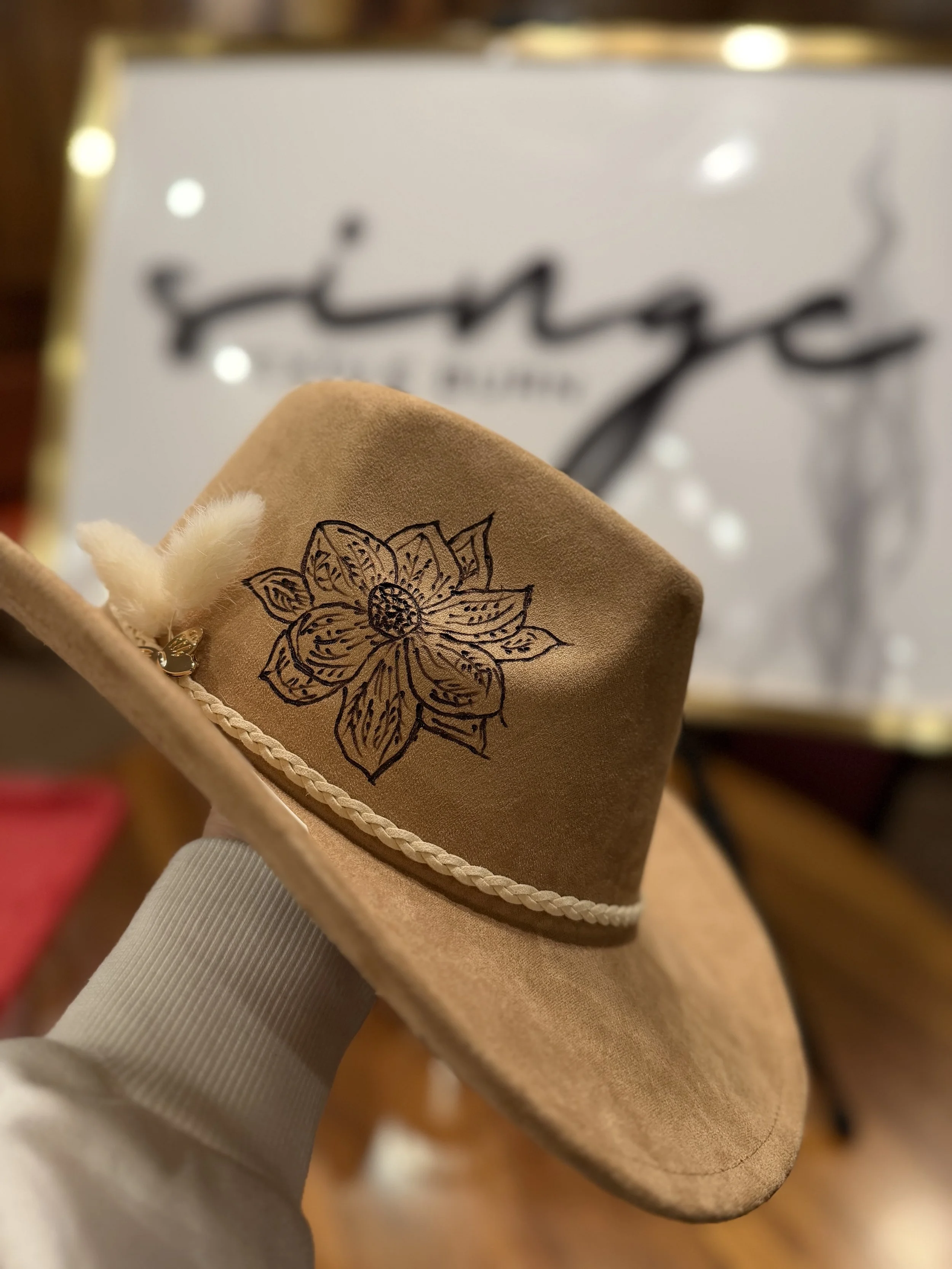 A beige suede shoe with black floral embroidery on the side, along with a braided trim near the sole, held by a hand with a white glove.