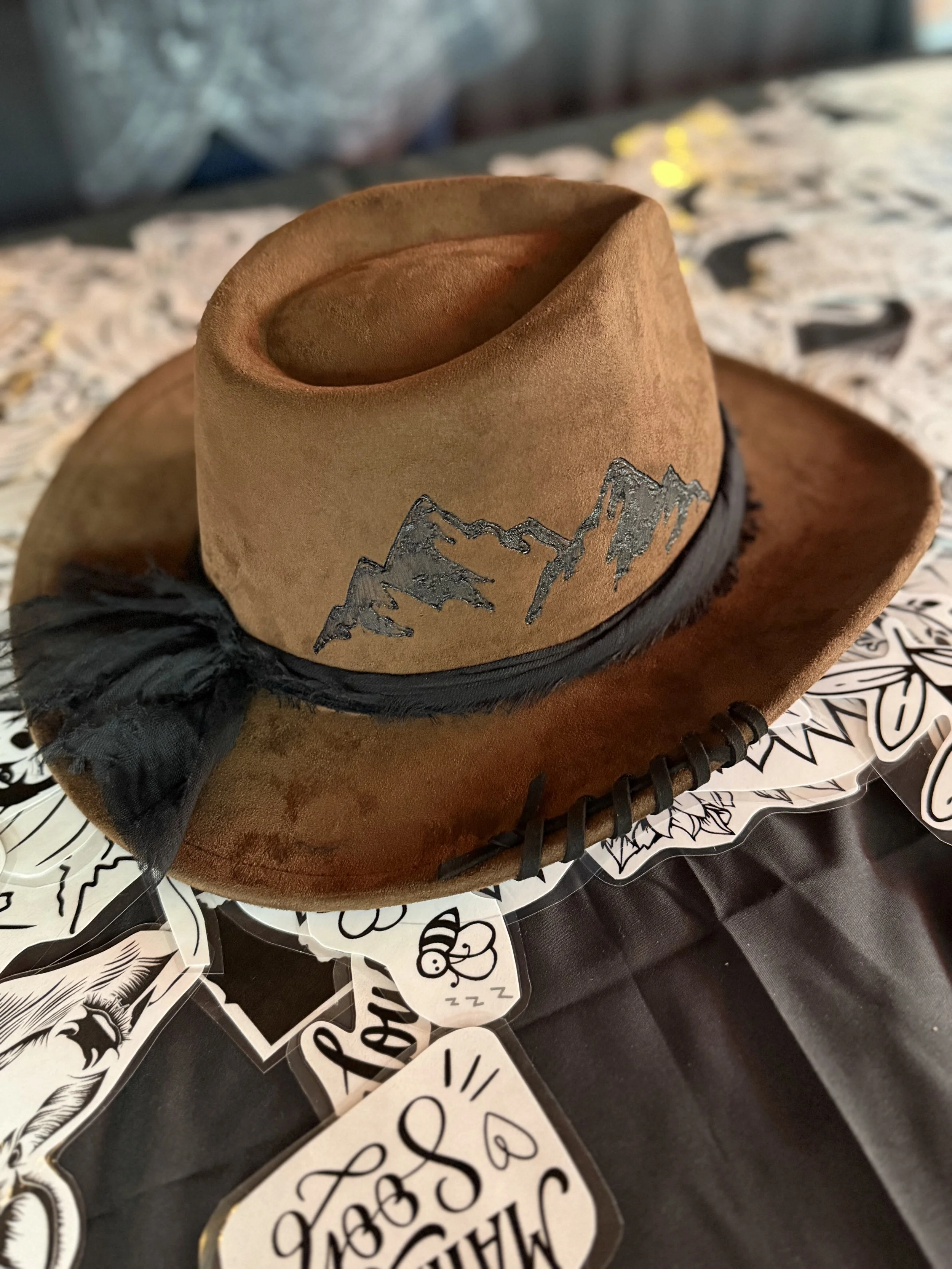 A brown cowboy hat with a mountain design, resting on a black surface surrounded by black and white stickers with various drawings and text, including a sleeping face with 'zzz' and a face with glasses.