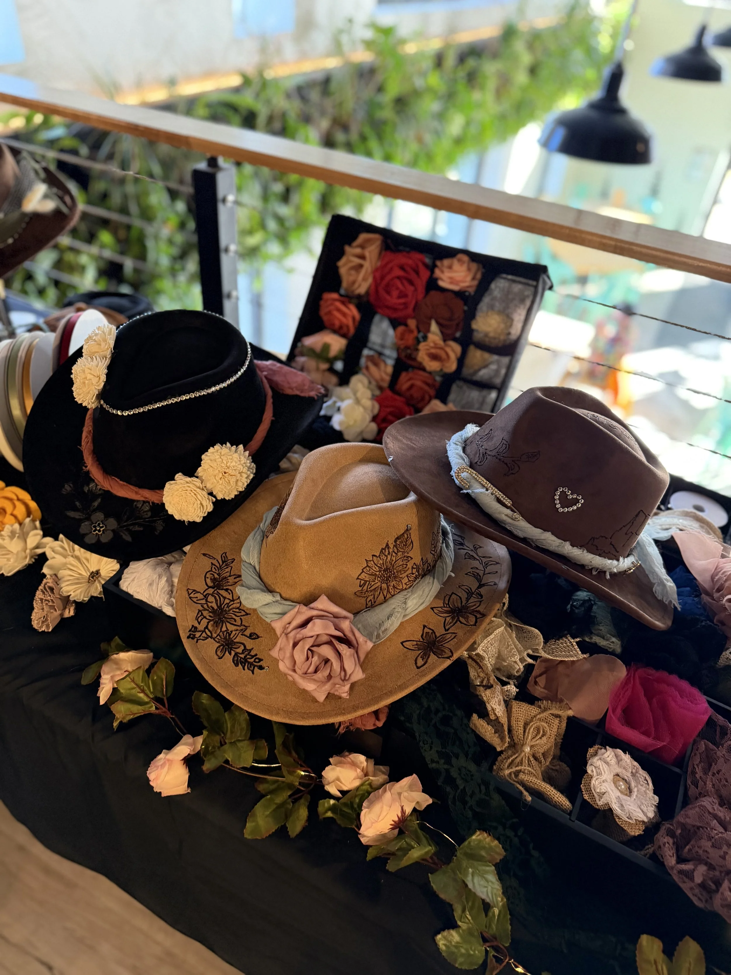 A display of various hats decorated with flowers and embroidery, arranged on a black surface with a floral garland border. The background shows a blurred outdoor setting with greenery and hanging lights.