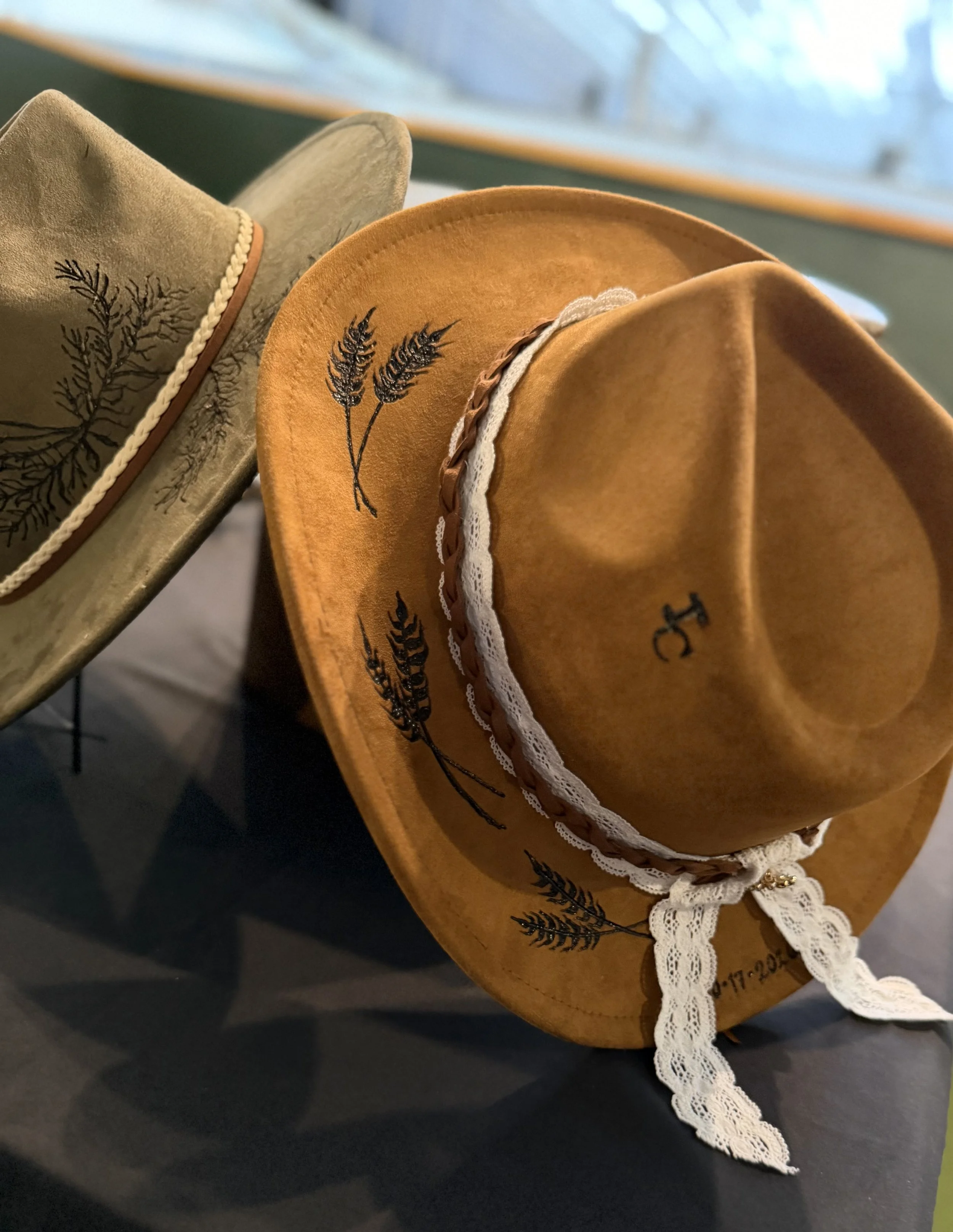 Close-up of two cowboy hats, one beige and one tan, decorated with embroidered black wheat design and white lace ribbon, resting on a dark surface.