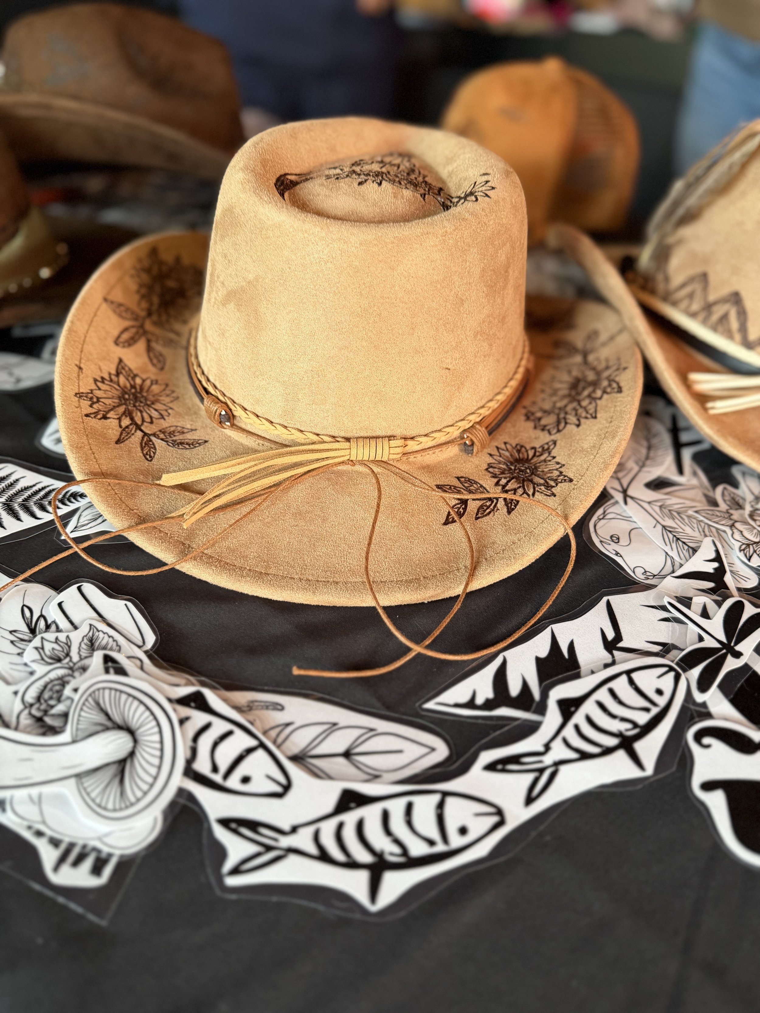 A light brown cowboy hat with floral and nature-themed black illustrations on the brim and crown, placed on a table surrounded by black and white stickers of fish and nature designs.