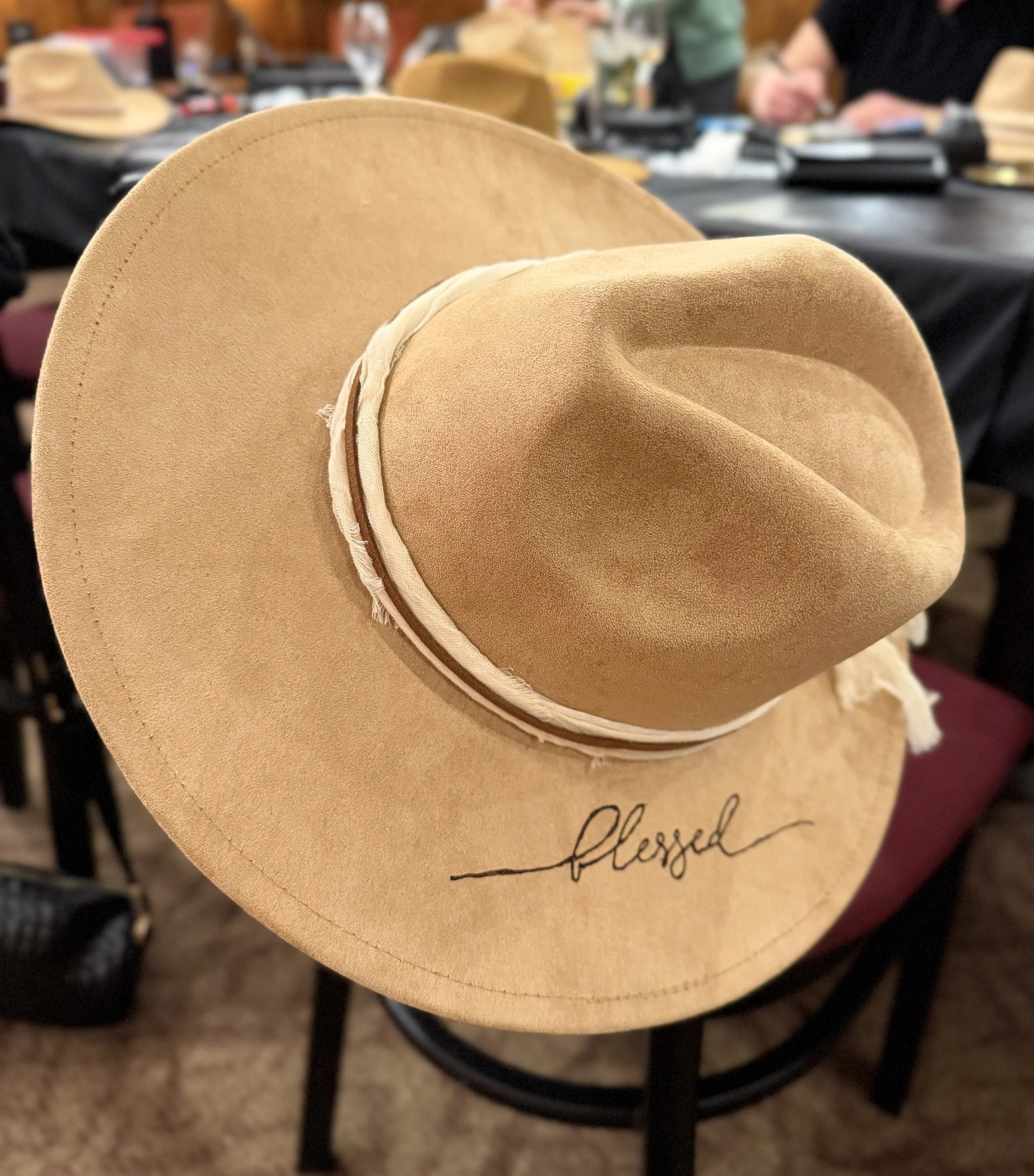 Close-up of a tan wide-brimmed felt hat with a beige band sitting on a black metal chair. The background shows other hats, tables, and people at a fair or market.