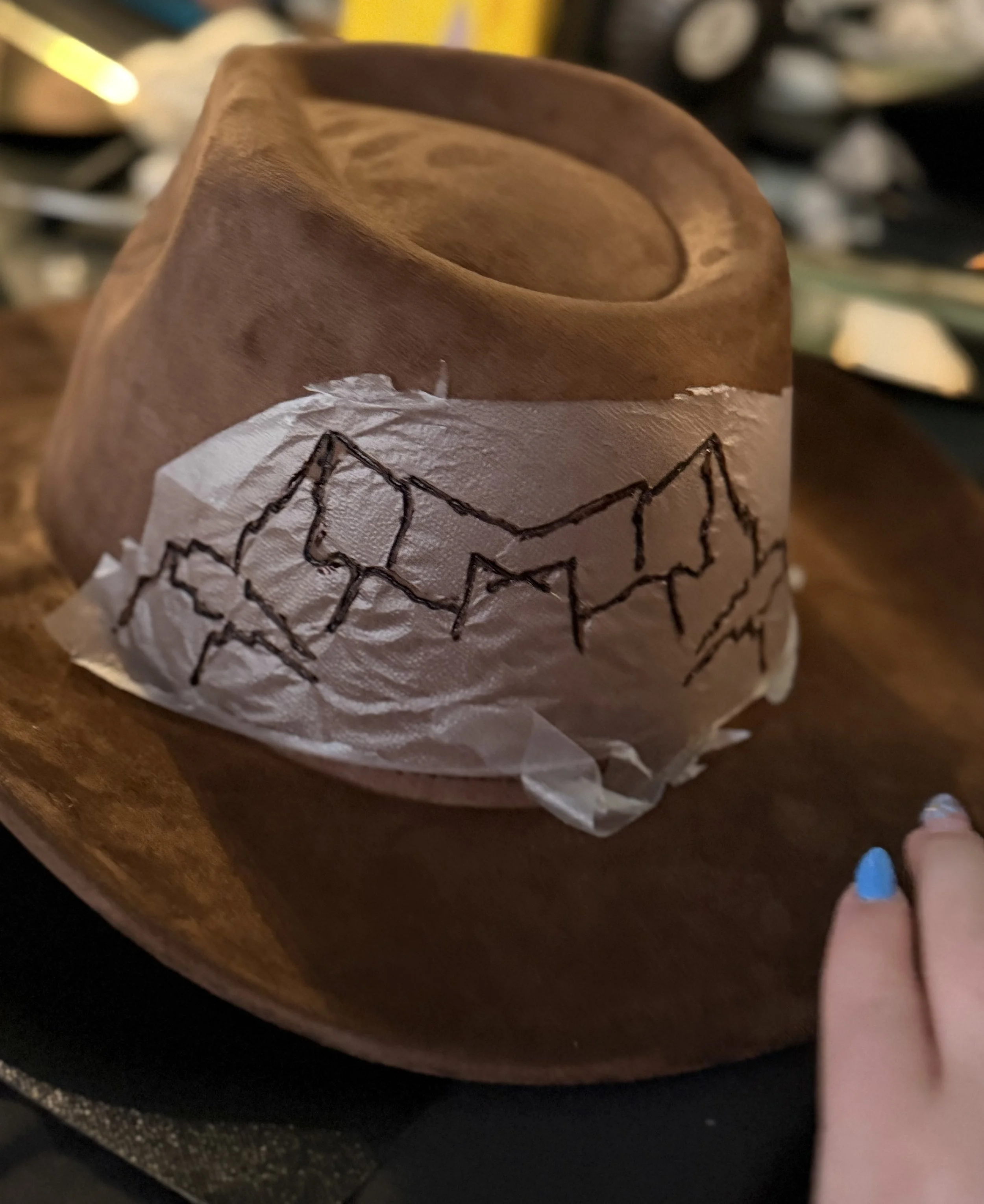 Close-up of a brown reusable coffee cup with a black illustration of a mountain range, sitting on a wooden surface, with a hand with blue painted nails holding the cup.