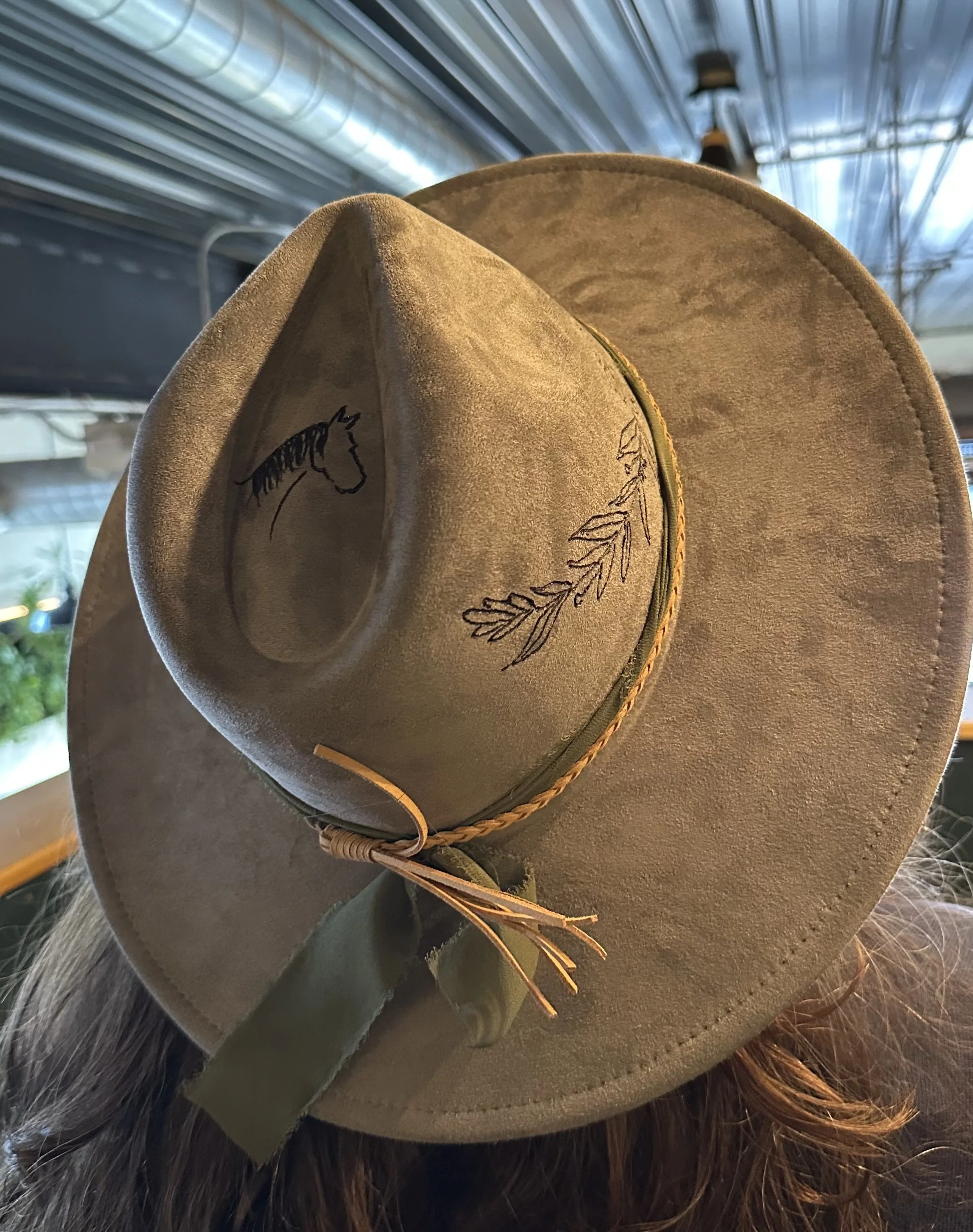 A tan cowboy hat with embroidered horse and leaf designs on the band, placed on a furry surface in a store or market.