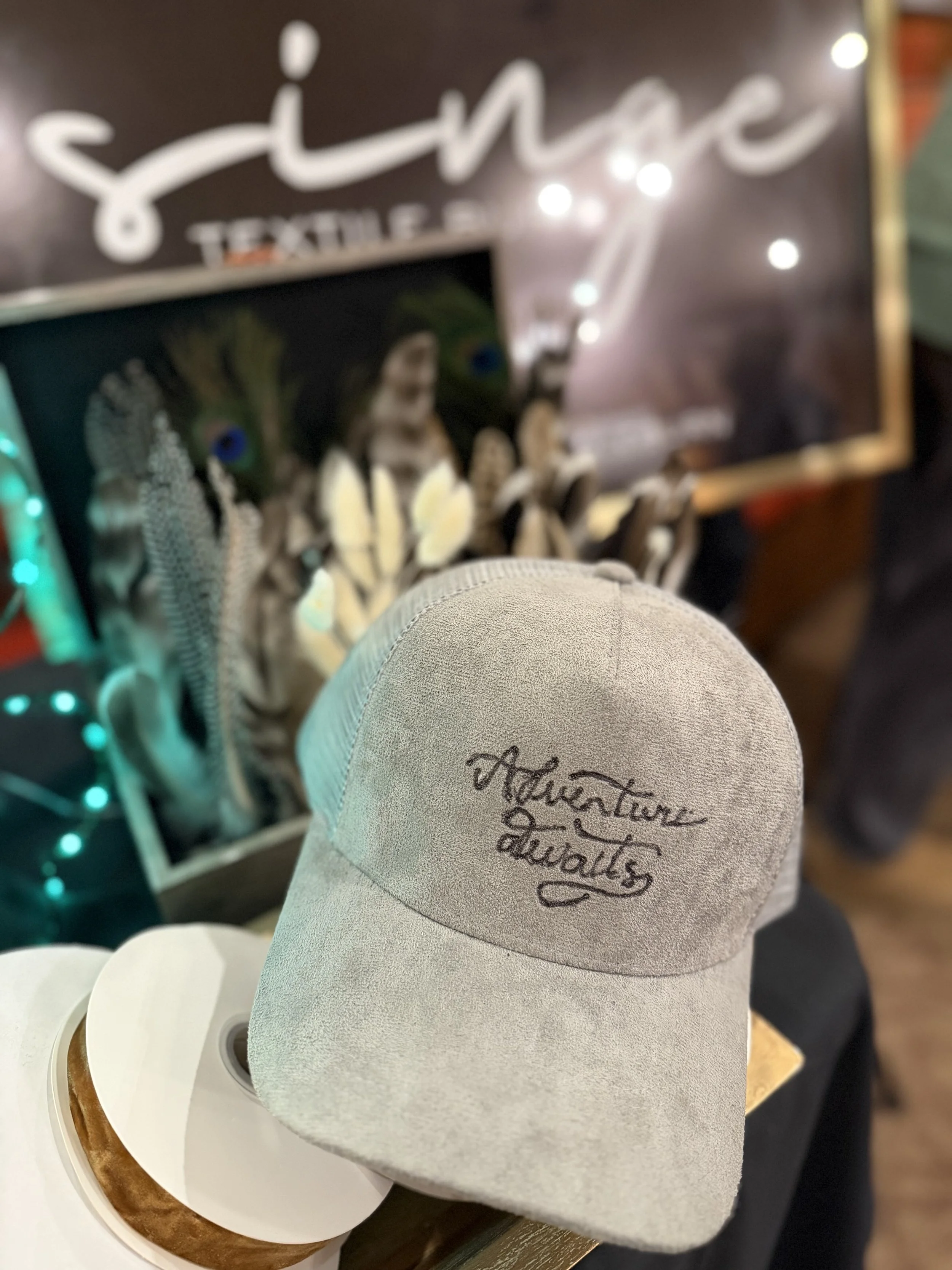 A white baseball cap with embroidered text that says "Finally an adult" resting on a table; blurred background showing decorative string lights and a sign that says "shh" and "TEXTILE STUDIO".