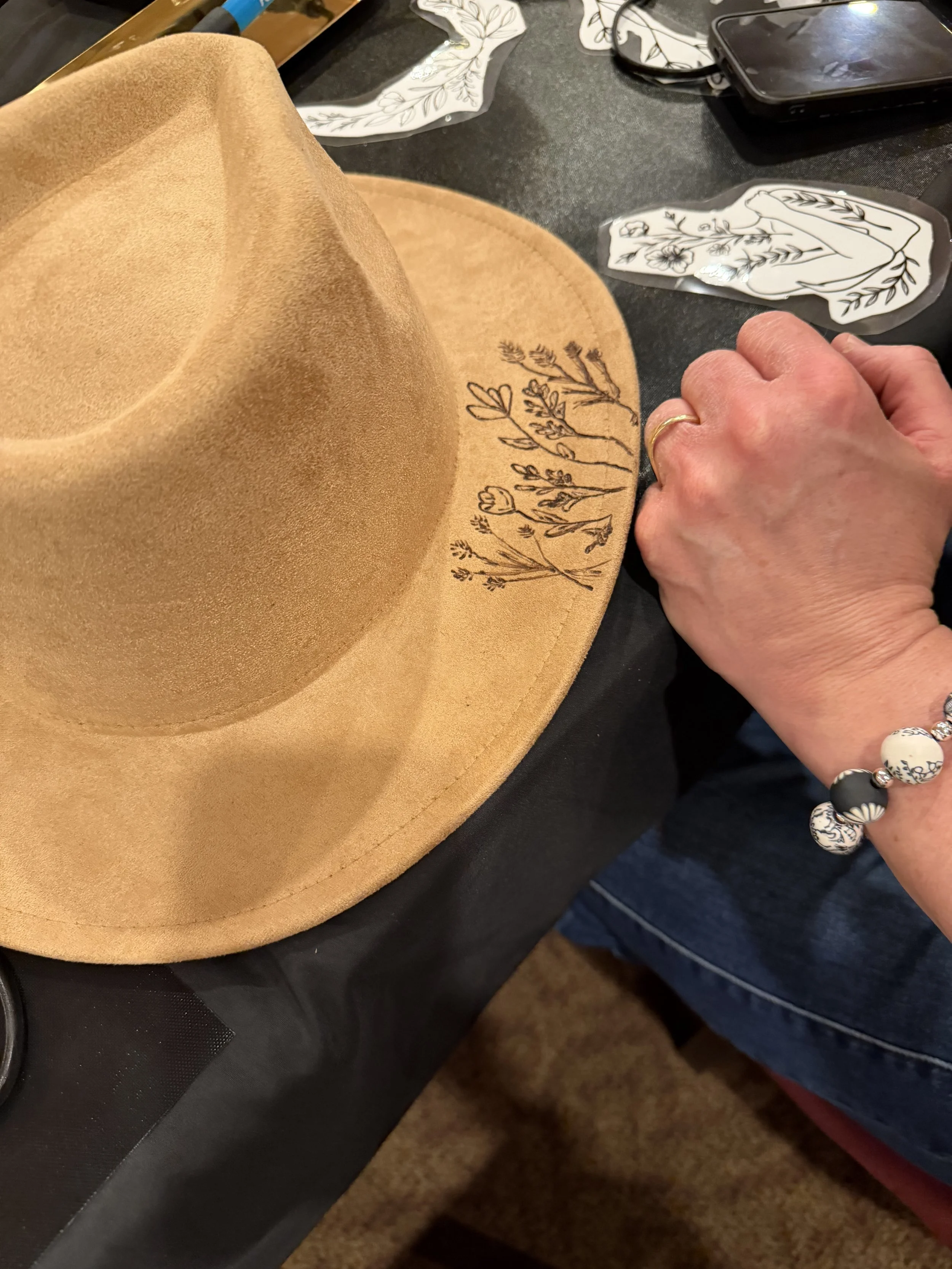 A tan wide-brimmed hat with black floral embroidery on the brim, placed on a black table, surrounded by a person's hand, a beaded bracelet, a phone, and decorative stickers of floral and abstract designs.