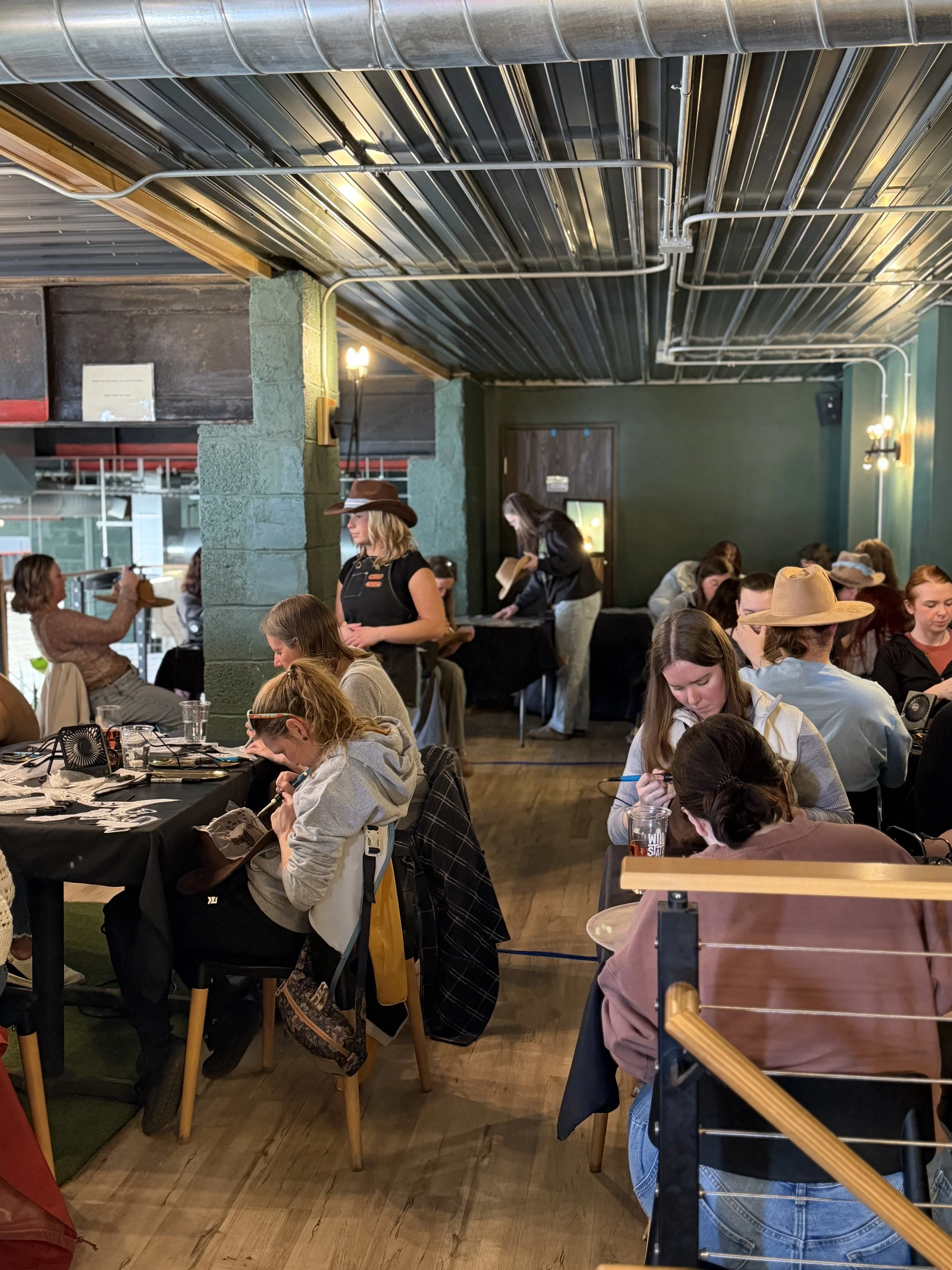 People sitting at tables in a restaurant or cafe, with some looking at their phones, others reading or talking, and staff serving food and taking orders. The ceiling is metallic with exposed pipes, and the walls are painted green.
