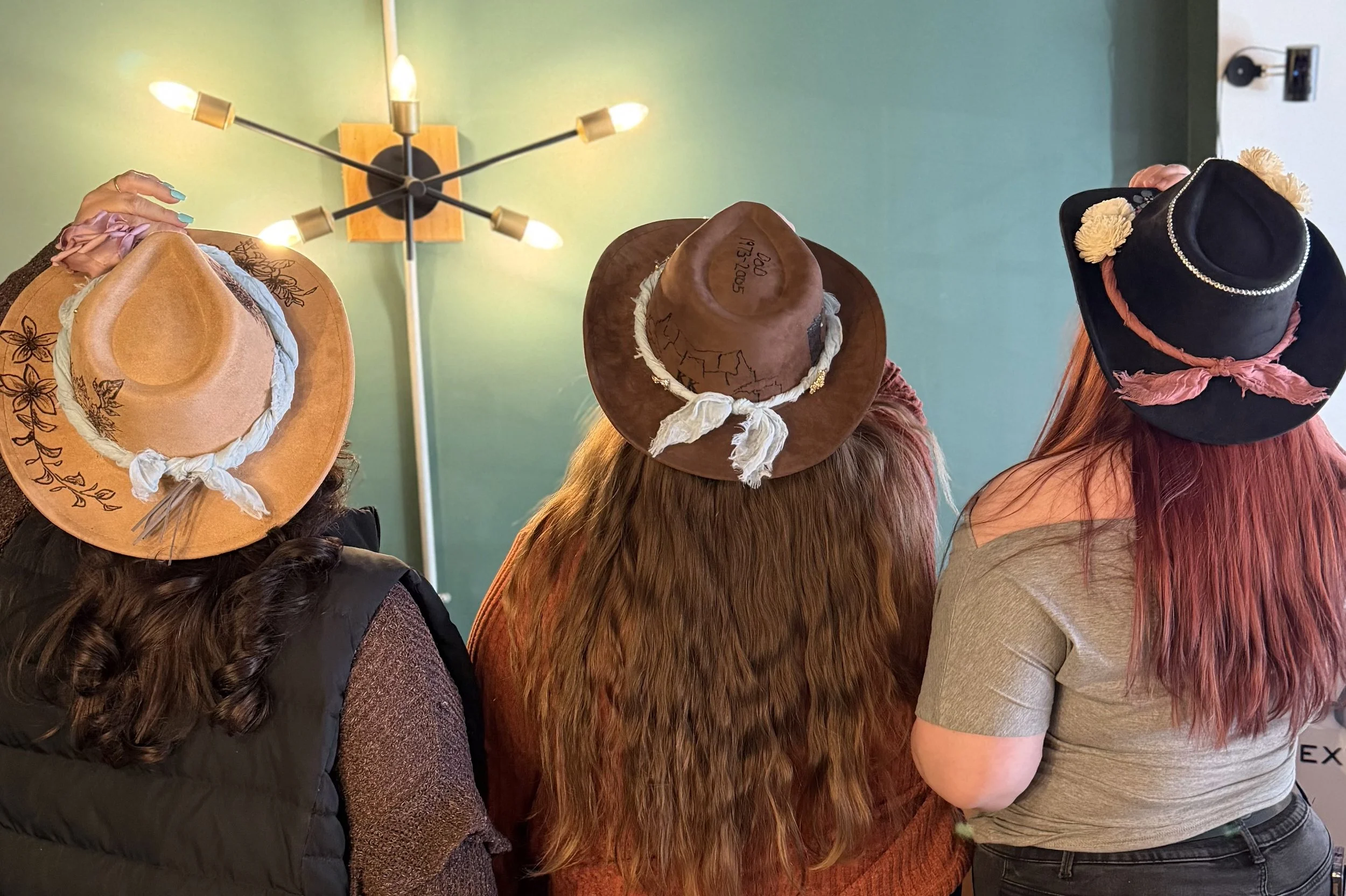 Three women wearing cowboy hats standing together indoors against a green wall, with a light fixture overhead.