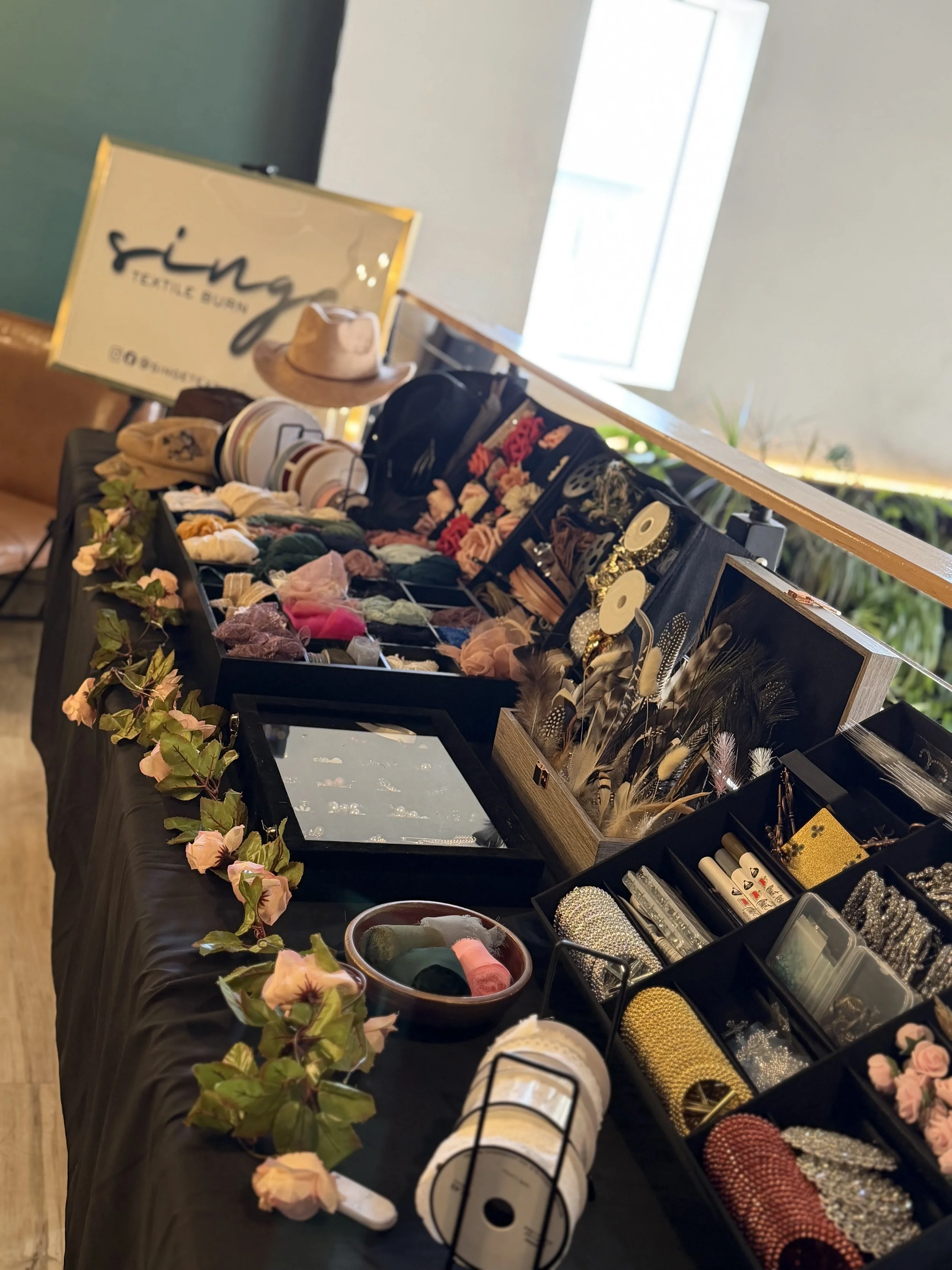 A table displaying various accessories and decorative items, including hats, hair accessories, jewelry, and fabric flowers, at a market or craft fair venue with a sign that says 'Jingles Textile Boutique'.