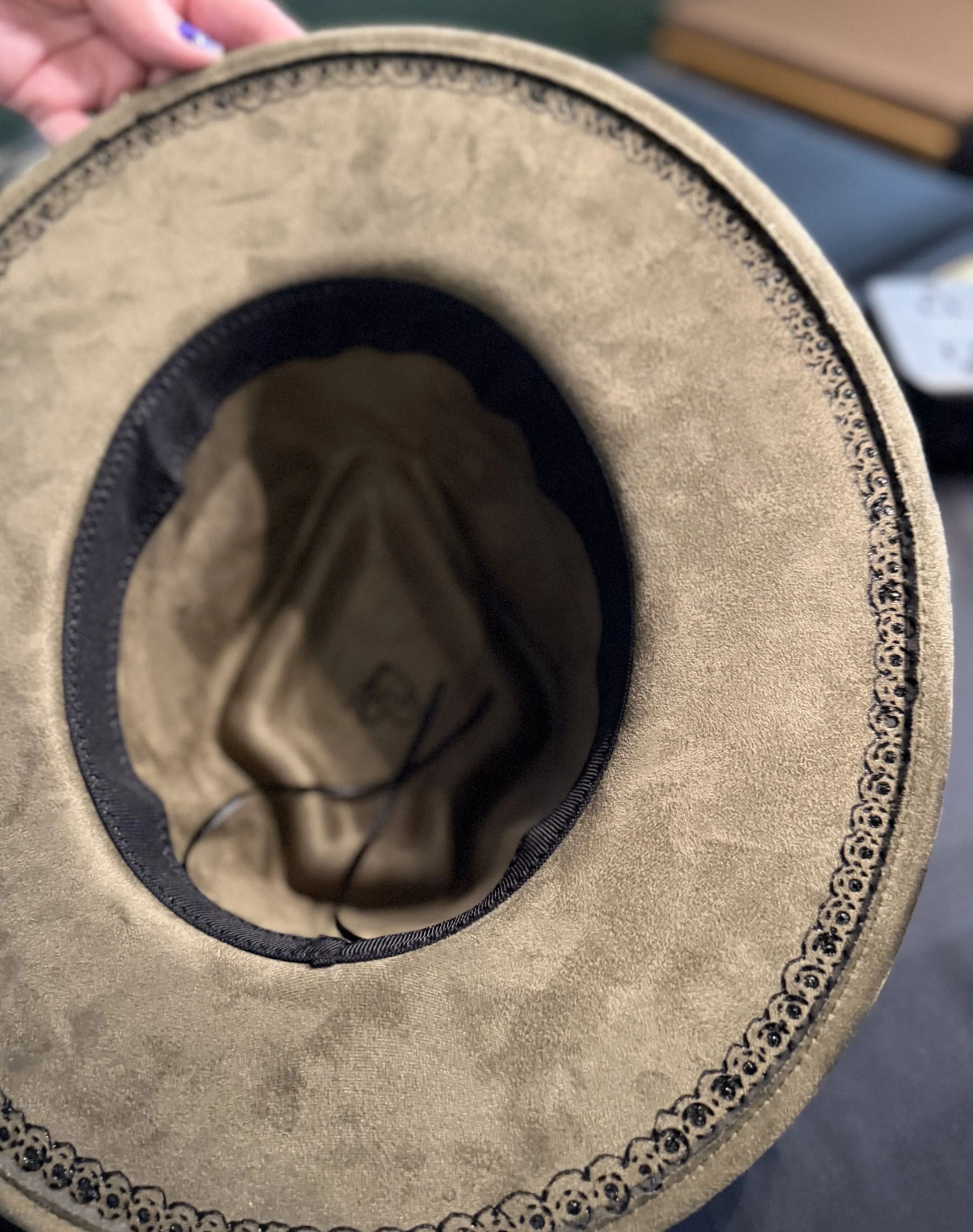 Top view of a beige hat with a black decorative edge and a black band inside, with a person's hand holding it from the top.