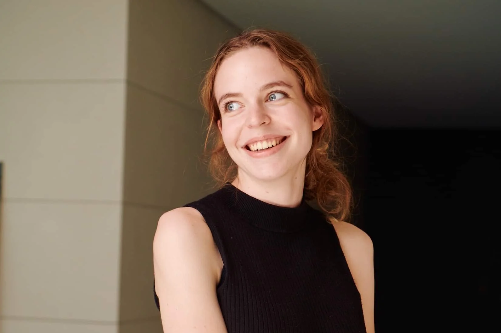 A young woman with red hair wearing a black sleeveless top smiling and looking to the side indoors.