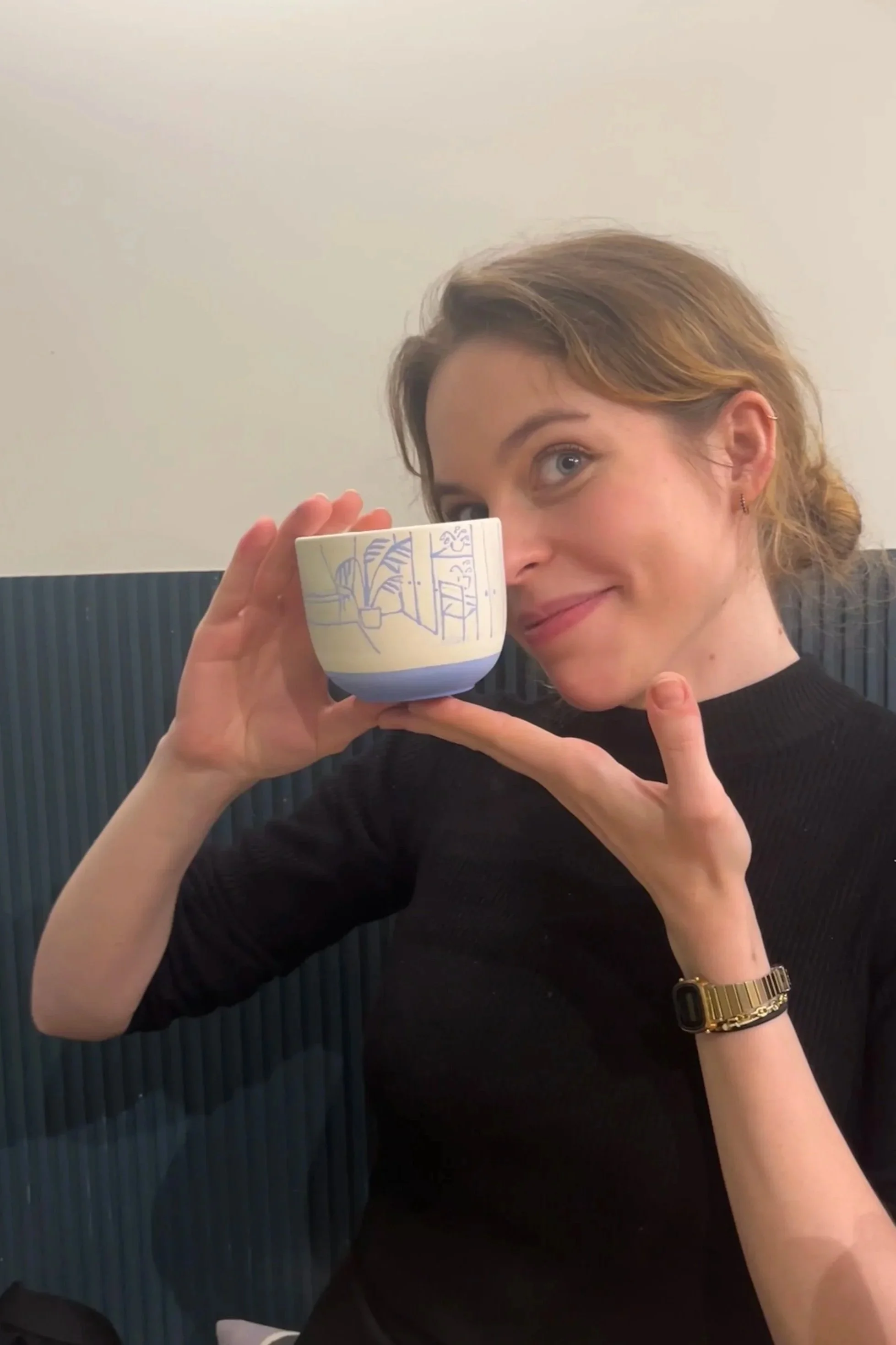 A woman with short, wavy hair and a black top holds a Japanese-style ceramic tea cup decorated with a blue line drawing. She is smiling and pointing at the cup, sitting against a dark blue, vertically striped wall.