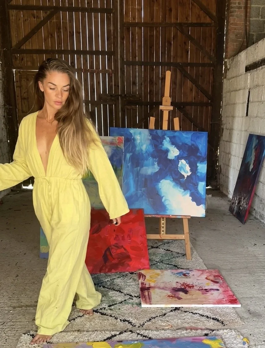 A woman wearing a bright yellow jumpsuit stands in an art studio with her eyes closed, surrounded by colorful abstract paintings on the floor and on an easel, with a rustic brick and wooden wall background.