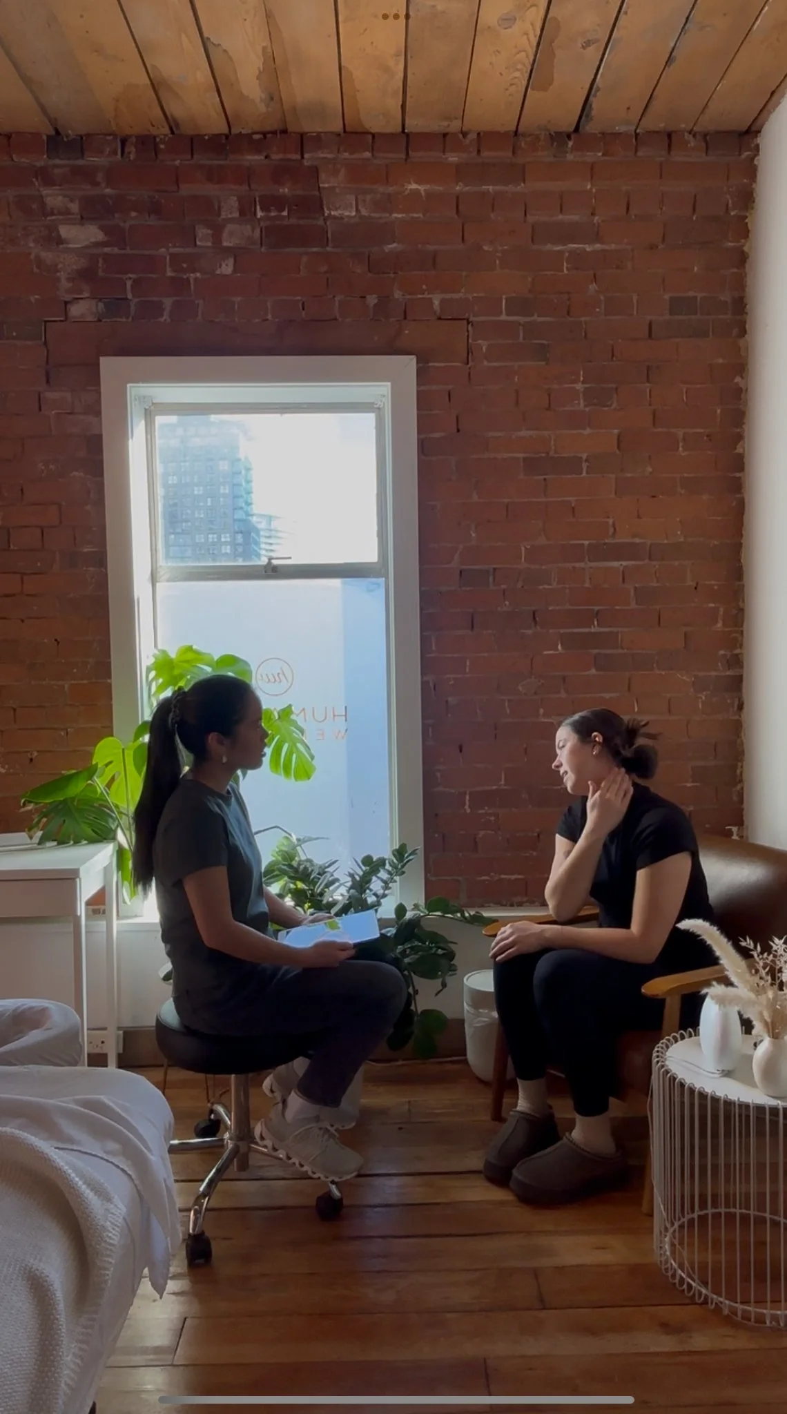 Massage therapist doing a patient intake in a cozy, natural light-filled room with an exposed brick wall, wooden ceiling, hardwood floor, potted plants, and minimalist décor.